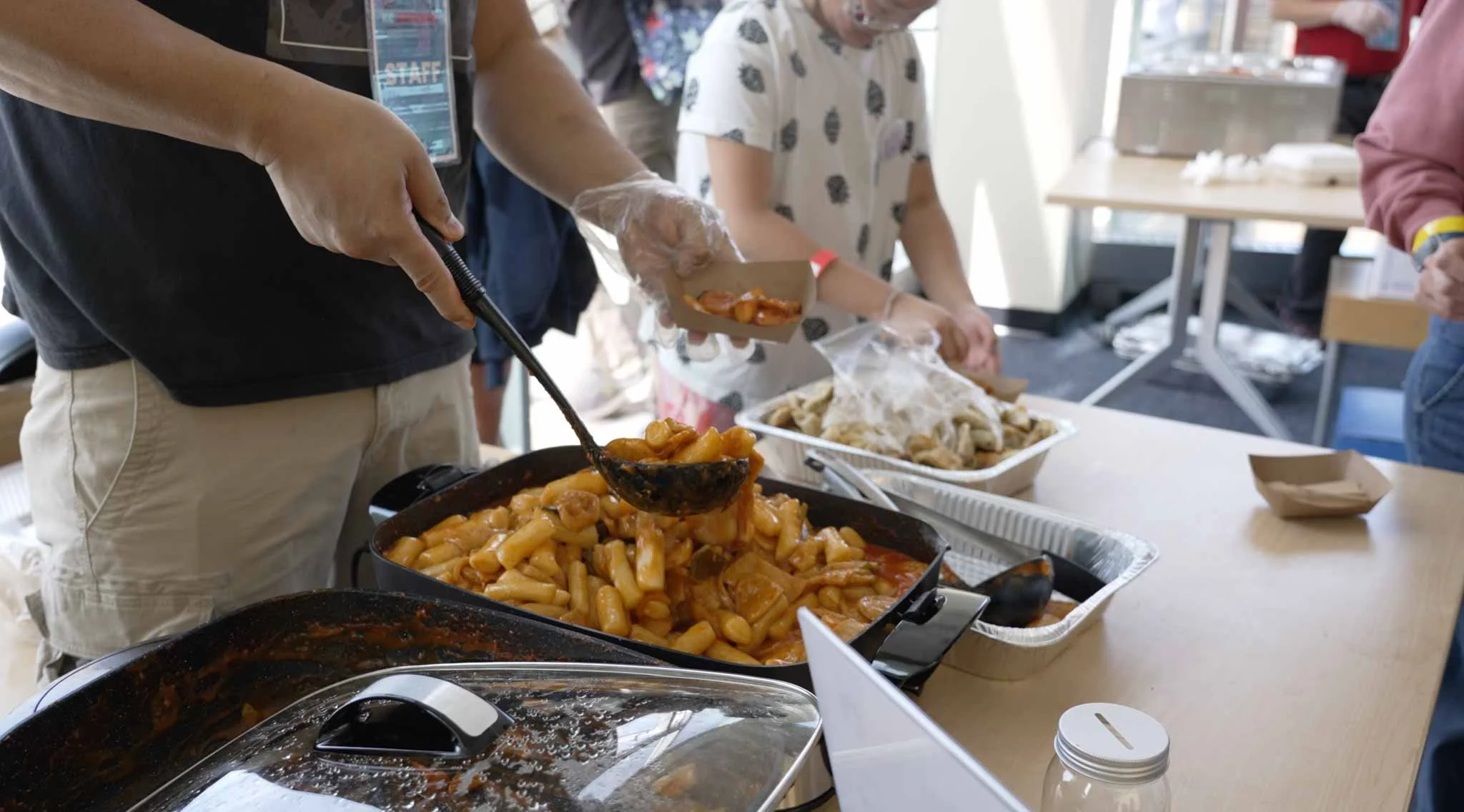 Person serving food from a large tray of baked pasta with sauce at a buffet-style meal, with other food trays and people in the background.