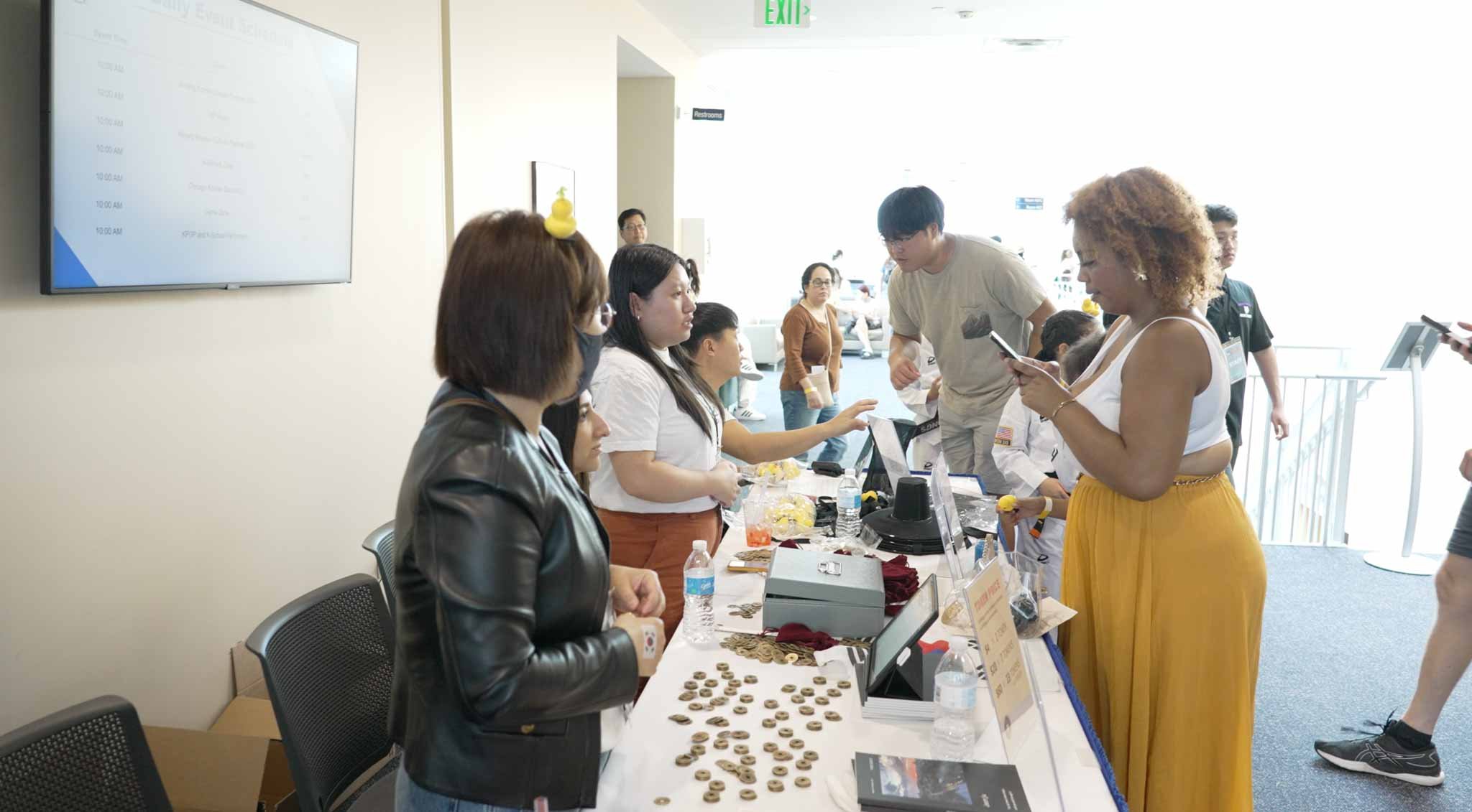 People at a registration or check-in table at an event, with a woman in yellow pants and a white top using her phone, and other attendees filling out papers or talking to staff.