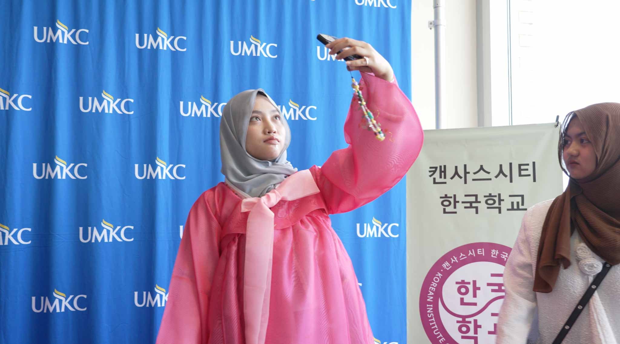 A young woman wearing a gray hijab and pink traditional Korean dress holding a smartphone with a beaded strap, standing in front of a blue background with 'UMKC' logos, and another woman wearing a brown hijab and beige blazer standing nearby.