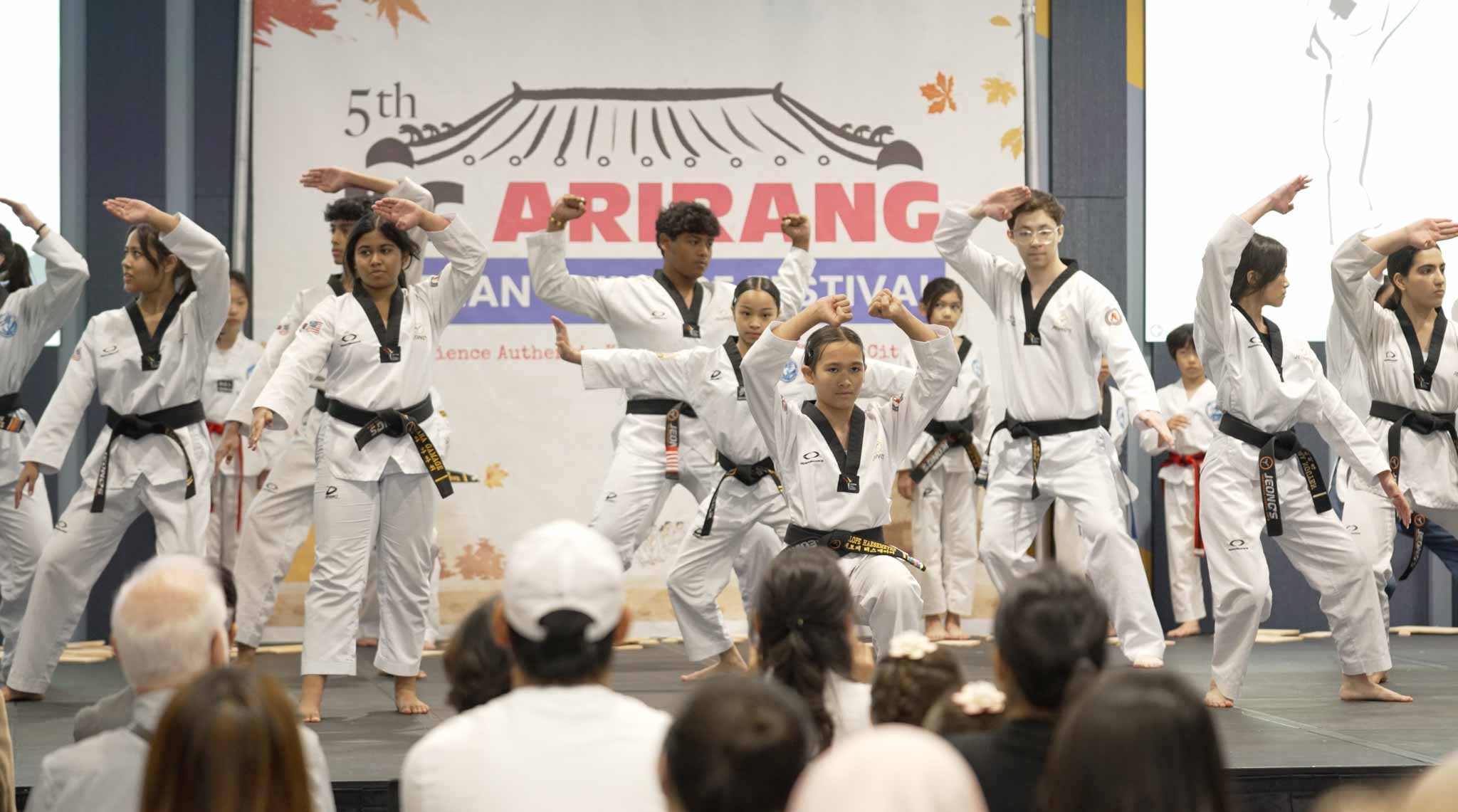 Group of young martial artists performing a Taekwondo demonstration on stage during the 5th Arirang Festival, with an audience watching.