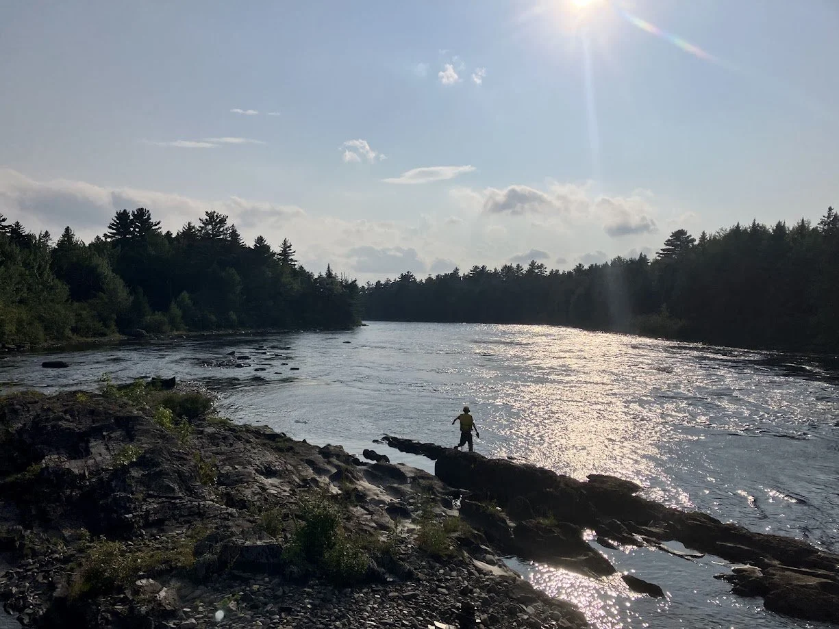 A person fishing on rocks along a river with trees on both sides, under a bright sky with the sun shining and some clouds.