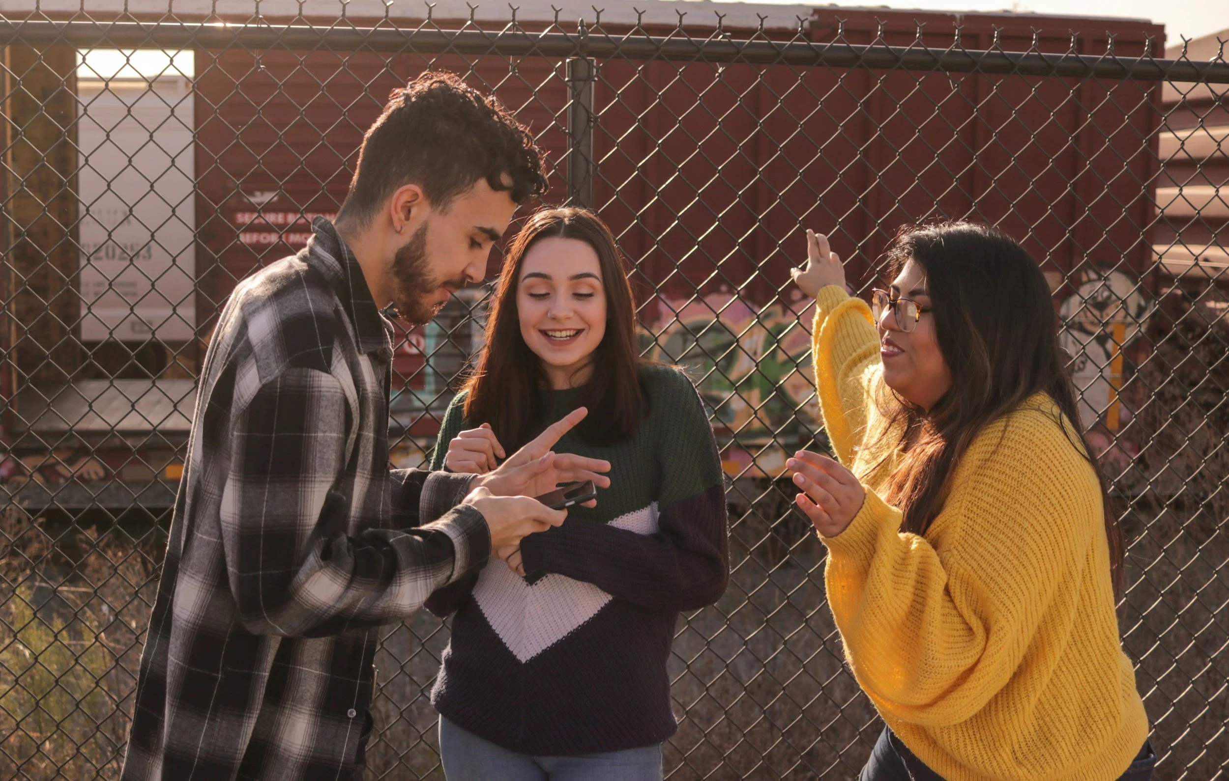 Three young adults, two women and one man, are standing behind a chain-link fence outdoors in warm sunlight. They are smiling and engaging happily, with the man showing something on a phone to the woman in the middle, who is pointing at it. The woman on the right is holding onto the fence and smiling.
