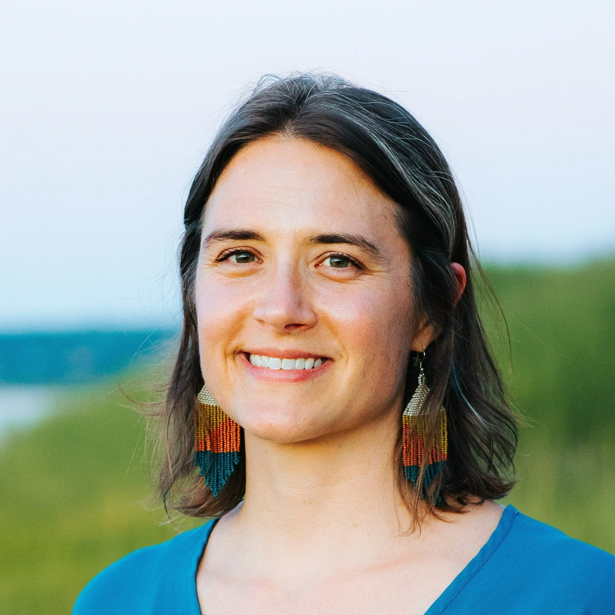 A woman with shoulder-length dark hair smiling outdoors, wearing colorful beaded earrings and a blue top, with a blurred natural landscape in the background.