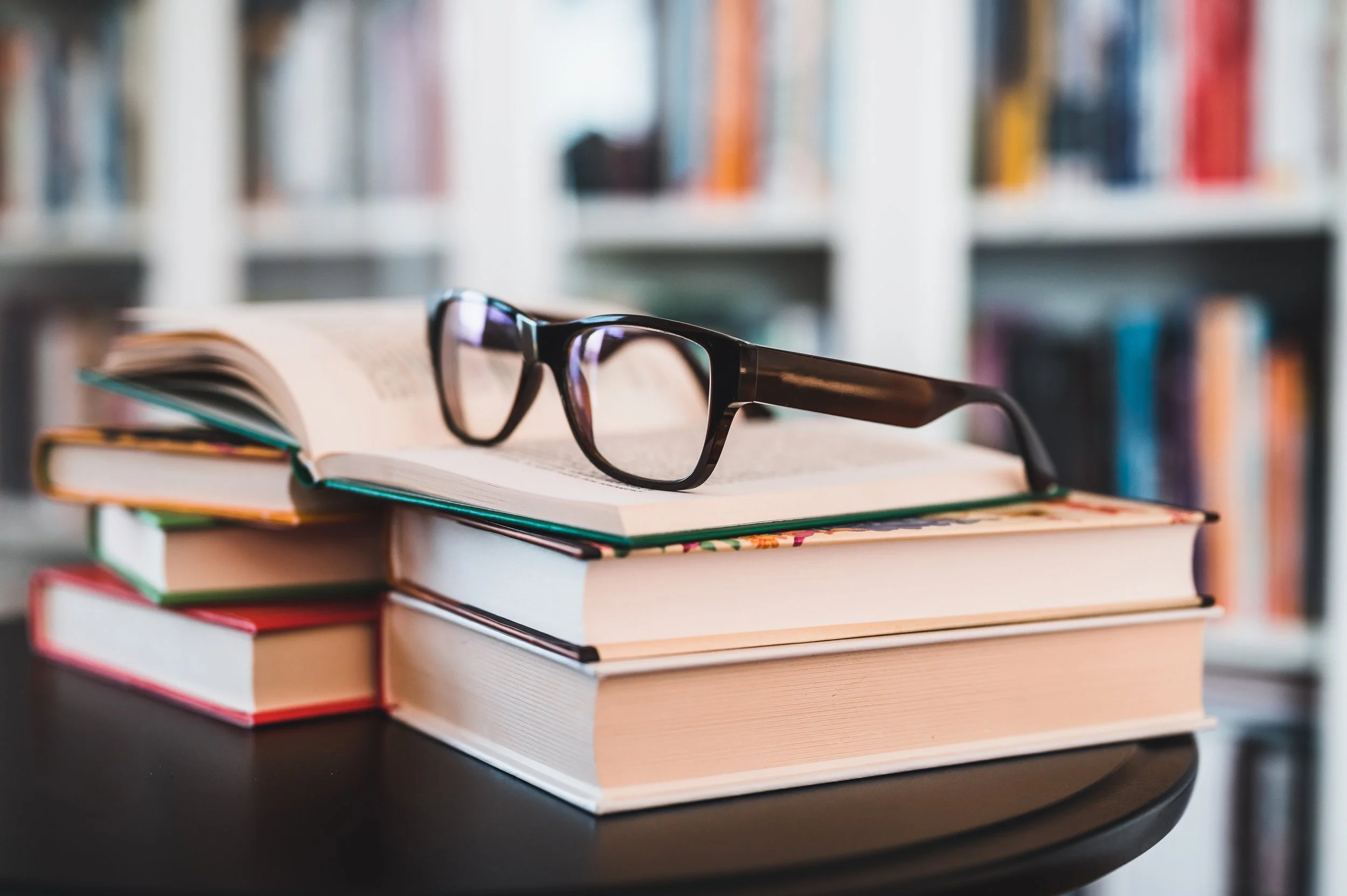 A stack of books with glasses resting on top, placed on a dark round table in a library setting with bookshelves in the background.