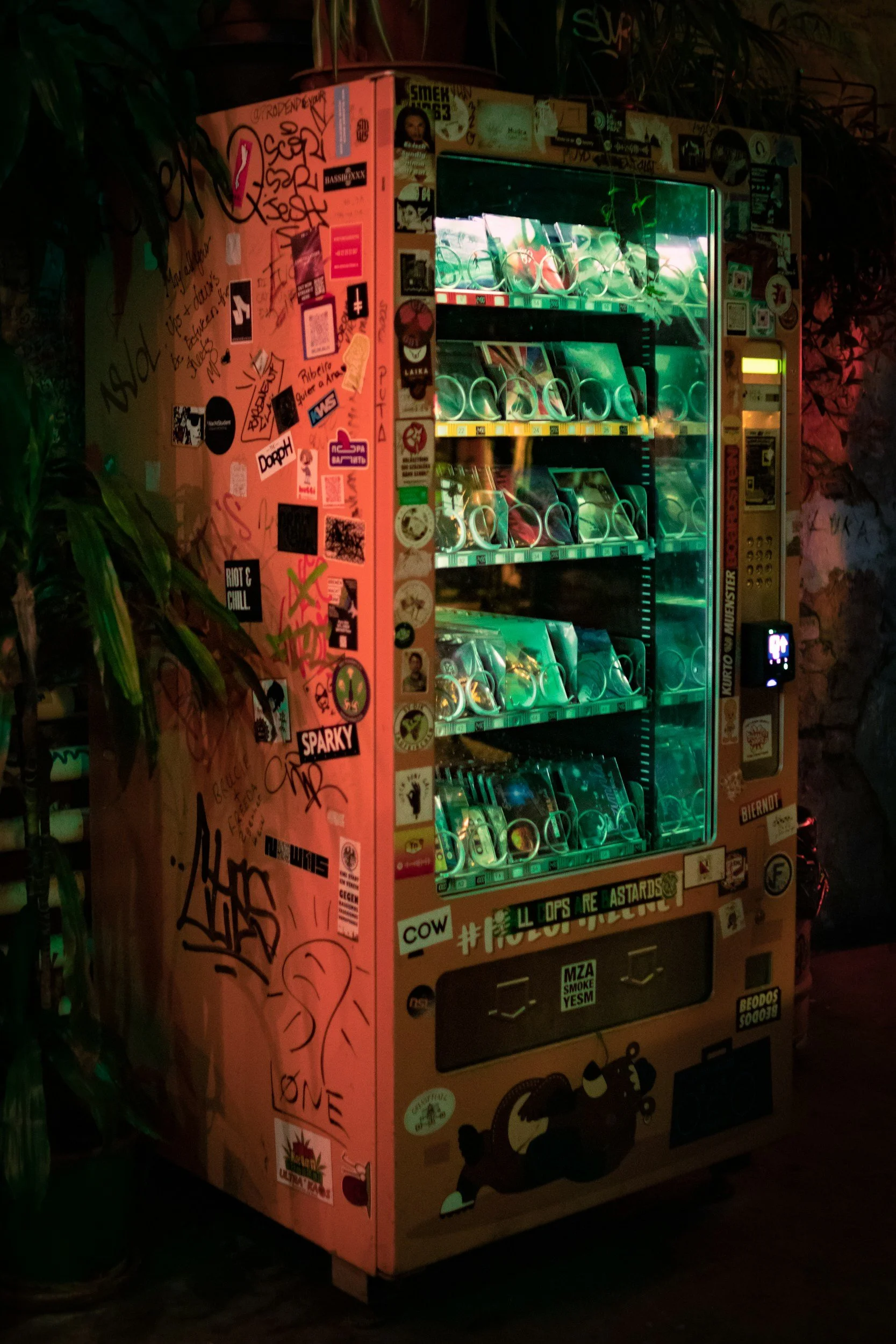 Een kleurrijke frisdrankautomaat met graffiti en stickers op de zijkanten, geplaatst in een gezellige, sfeervolle omgeving met planten en stenen muren.