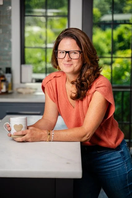 Woman with brown hair and glasses sitting at a kitchen counter, holding a white mug with a heart design, in front of a window with green trees outside.