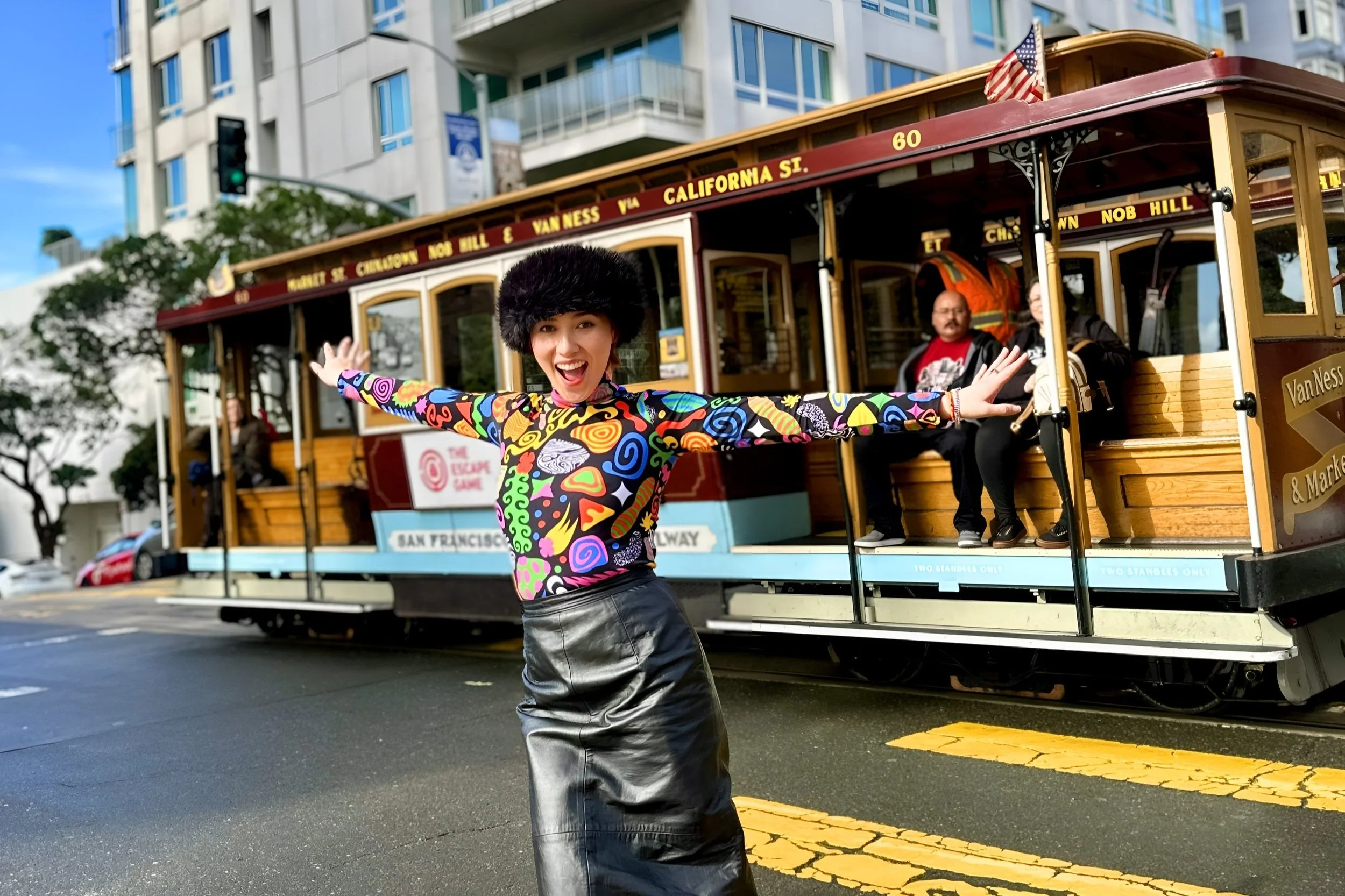 A woman wearing a colorful longsleeve top and a leather skirt poses in front of a Cable Car passing by at a street intersection in San Francisco