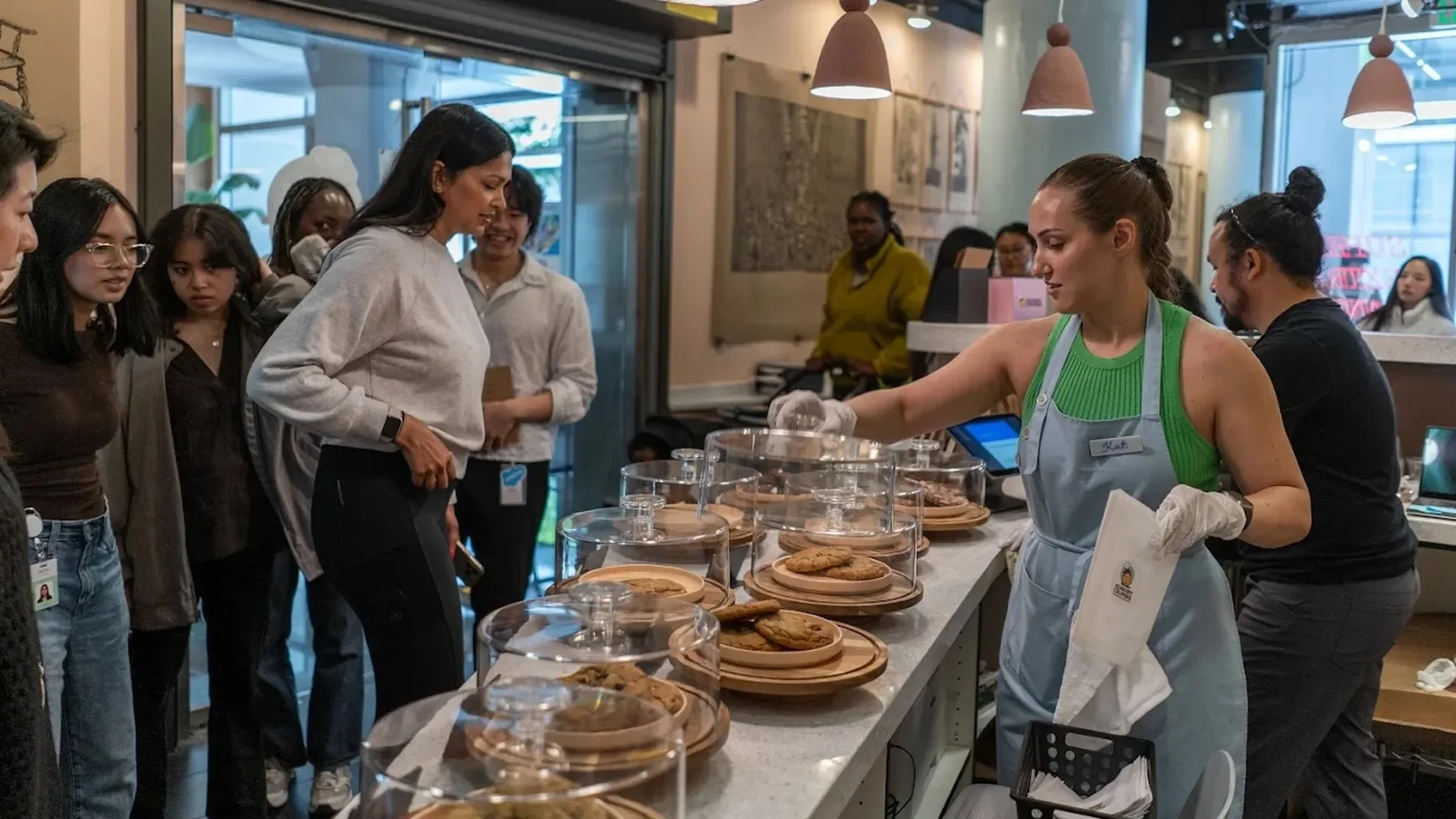 Customers line up at a cookie shop counter as a staff member in an apron serves fresh cookies from display trays.