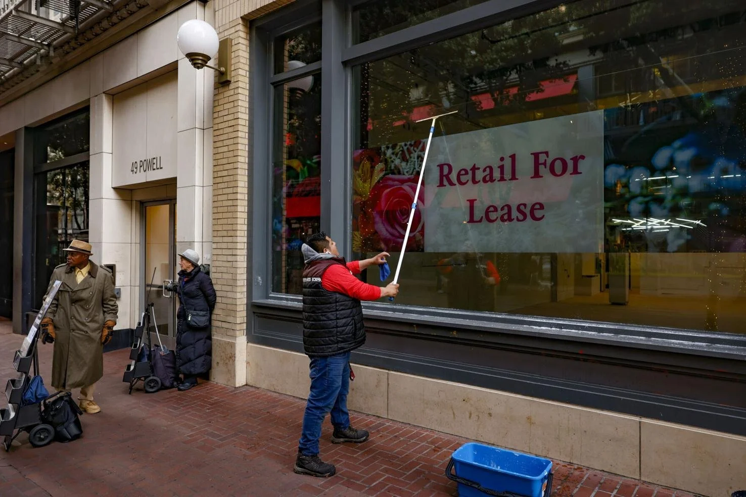 A man cleans the window of a vacant storefront in San Francisco with a large "Retail For Lease" sign, while two people stand nearby.