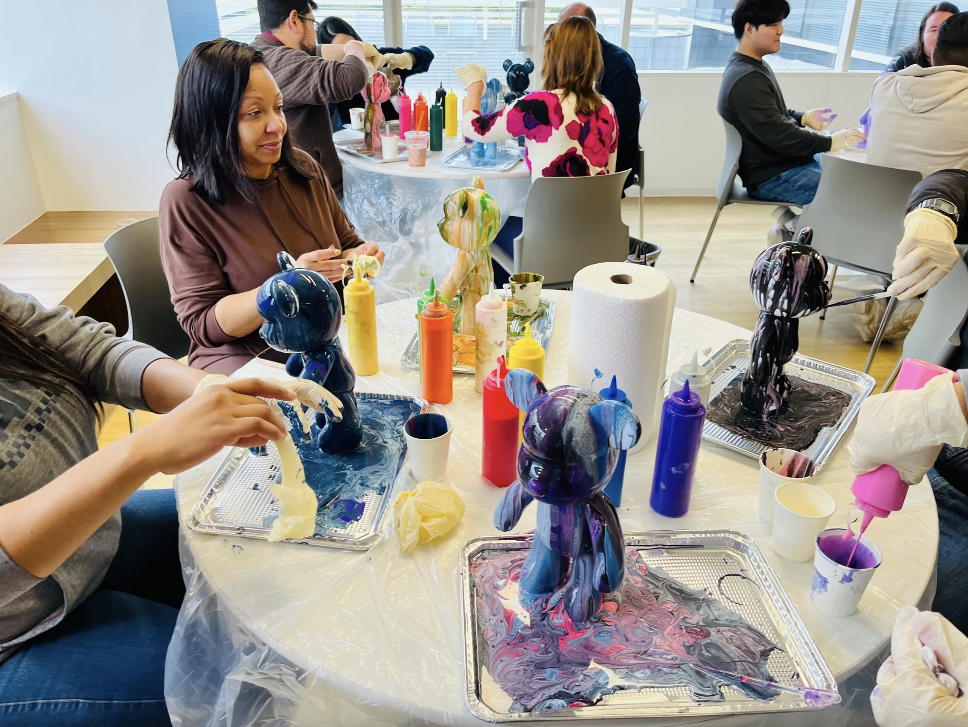 Groups of diverse people sit at tables and pour different colored paint on figurines