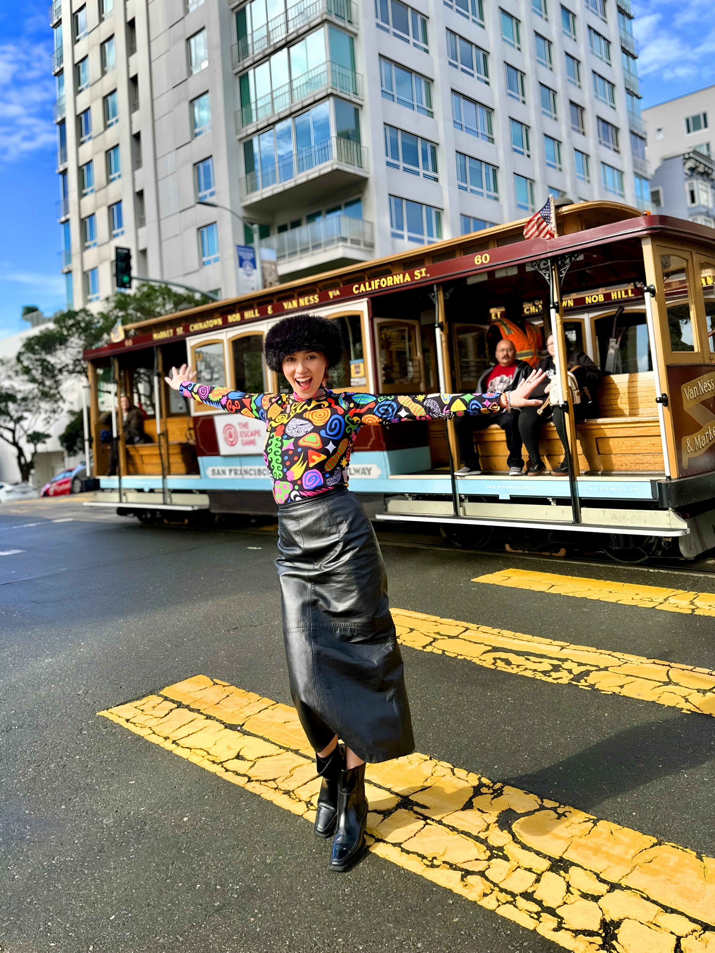 A woman wearing a colorful top and leather skirt poses in front of a cable car at a San Francisco street intersection