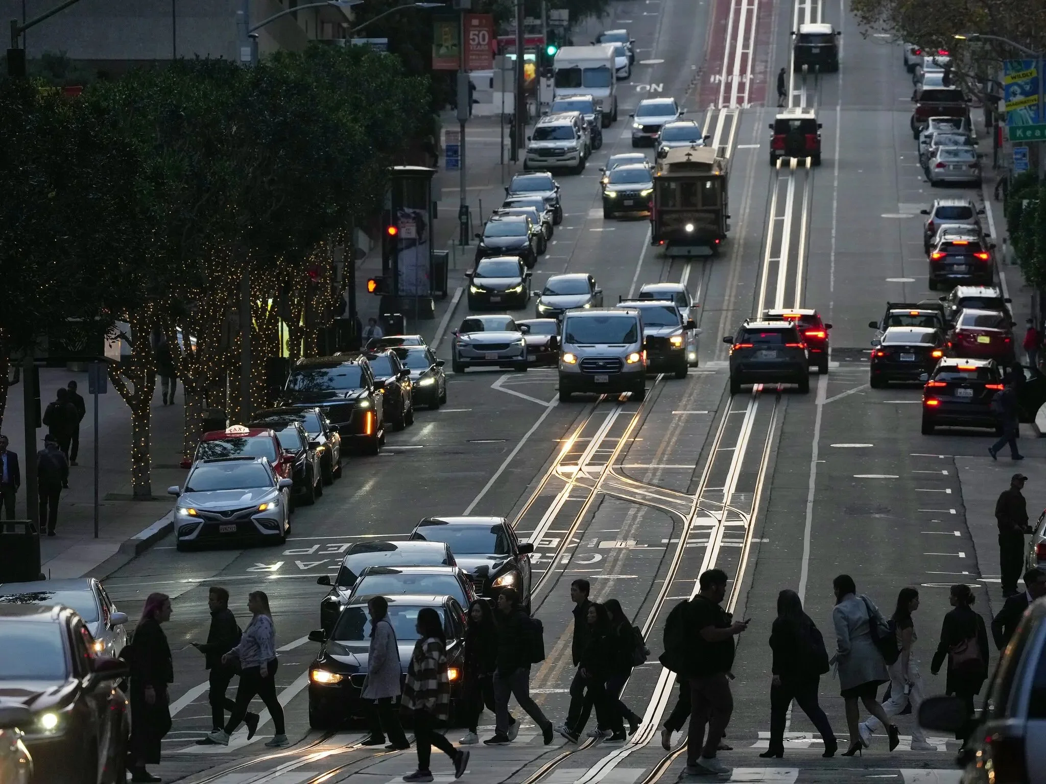 Pedestrians cross a bustling San Francisco street at dusk as cars, buses, and a cable car navigate the traffic.