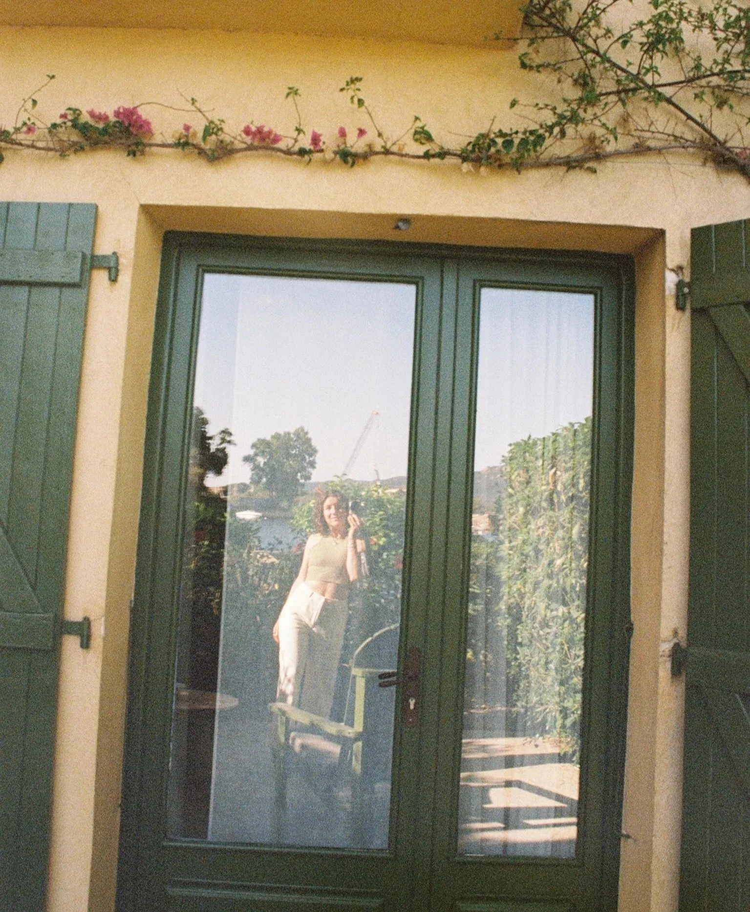 Reflection of a smiling woman in a glass door, holding a phone in her right hand, with a garden and trees in the background.