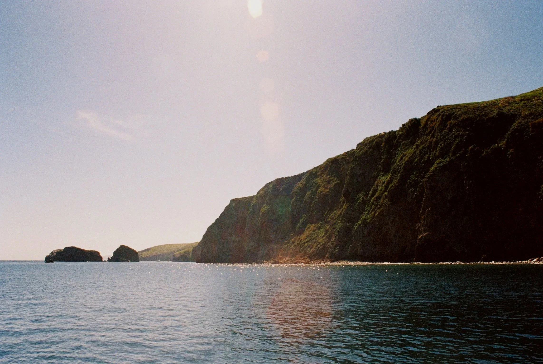 Coastal landscape with green cliffs rising from the water under a clear blue sky, with sunlight creating a lens flare.