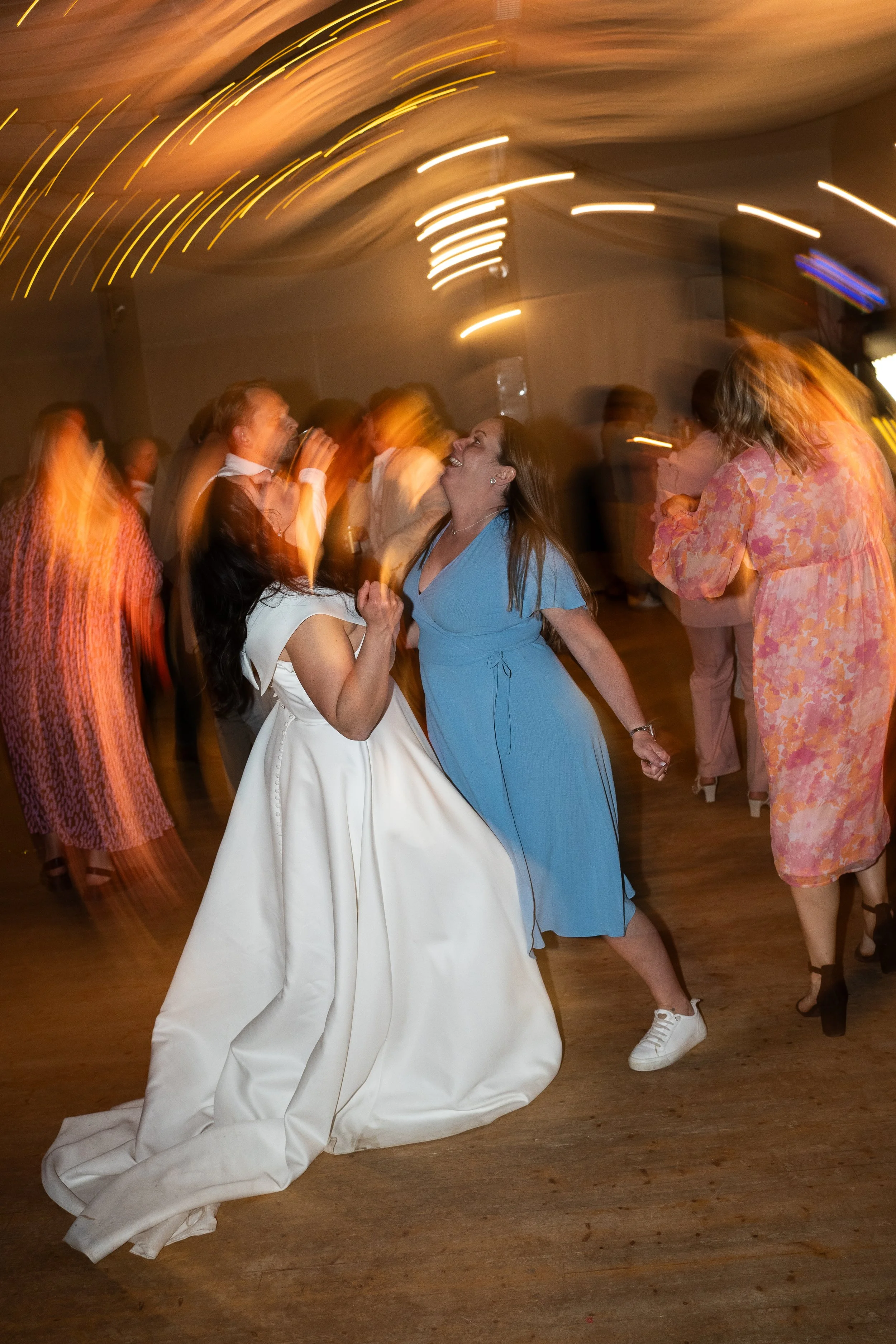 Två kvinnor dansar glatt tillsammans på dansgolvet, en i vit klänning och en i blå klänning, med andra personer omkring dem som dansar i en festlig miljö.