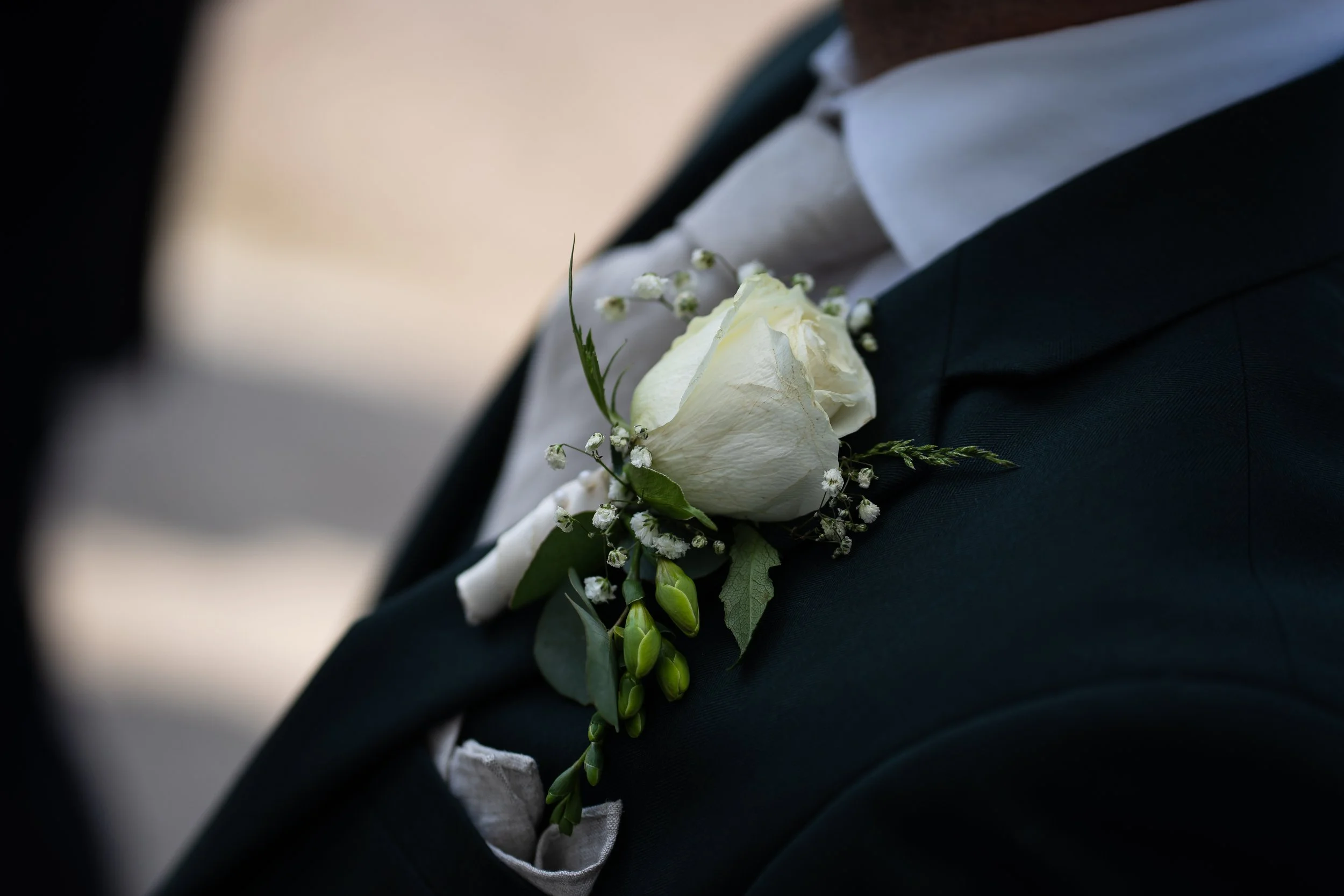 Brudgummen bär en vit blomsterboutonnière med en vit ros, gröna blad och små vita blommor på kavaj.