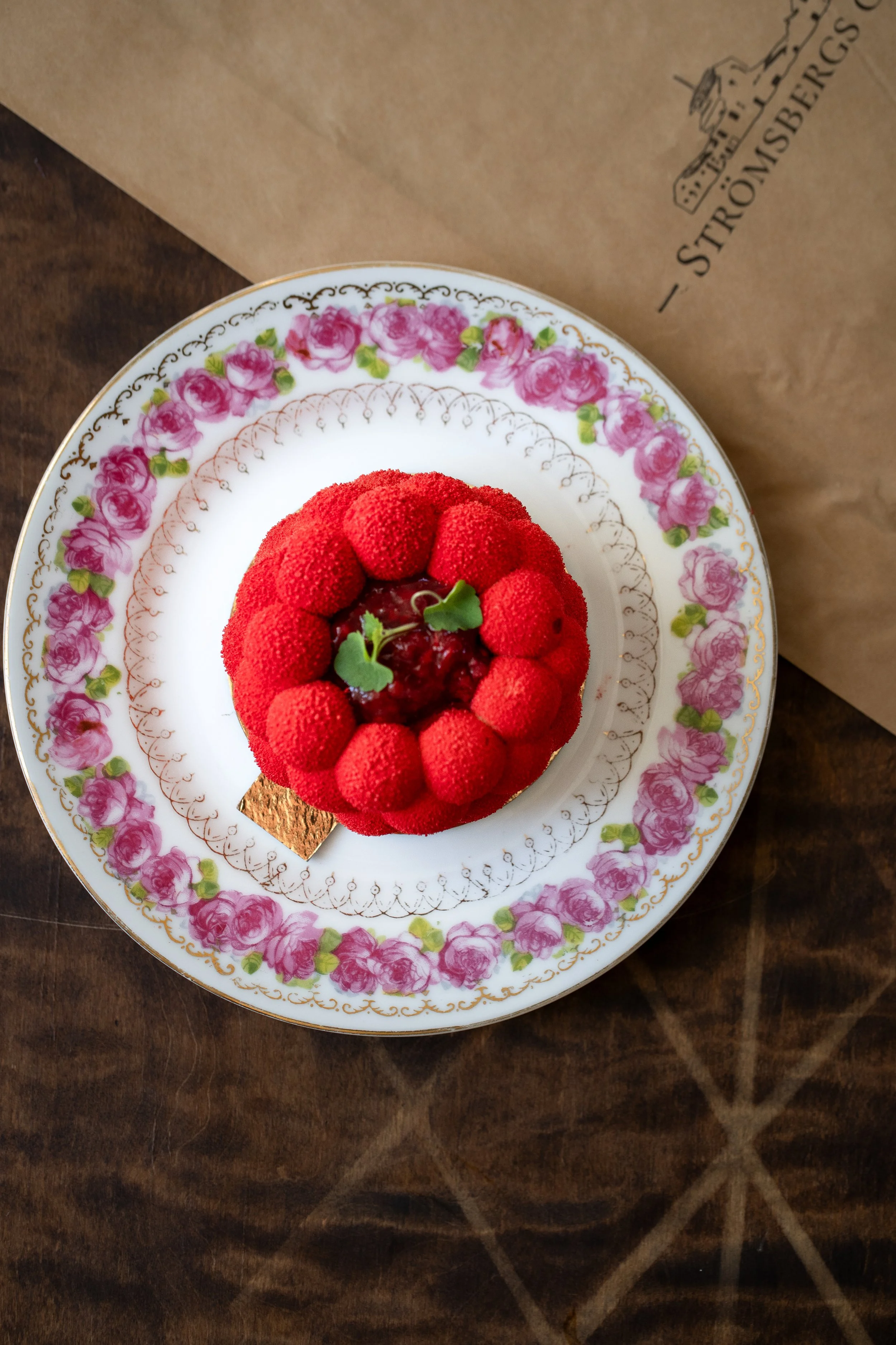 En tårtdekoration med röda bär och gröna örter på ett porslinsfat med rosa blommor och guldkant, placerad på ett mörkt träbord.