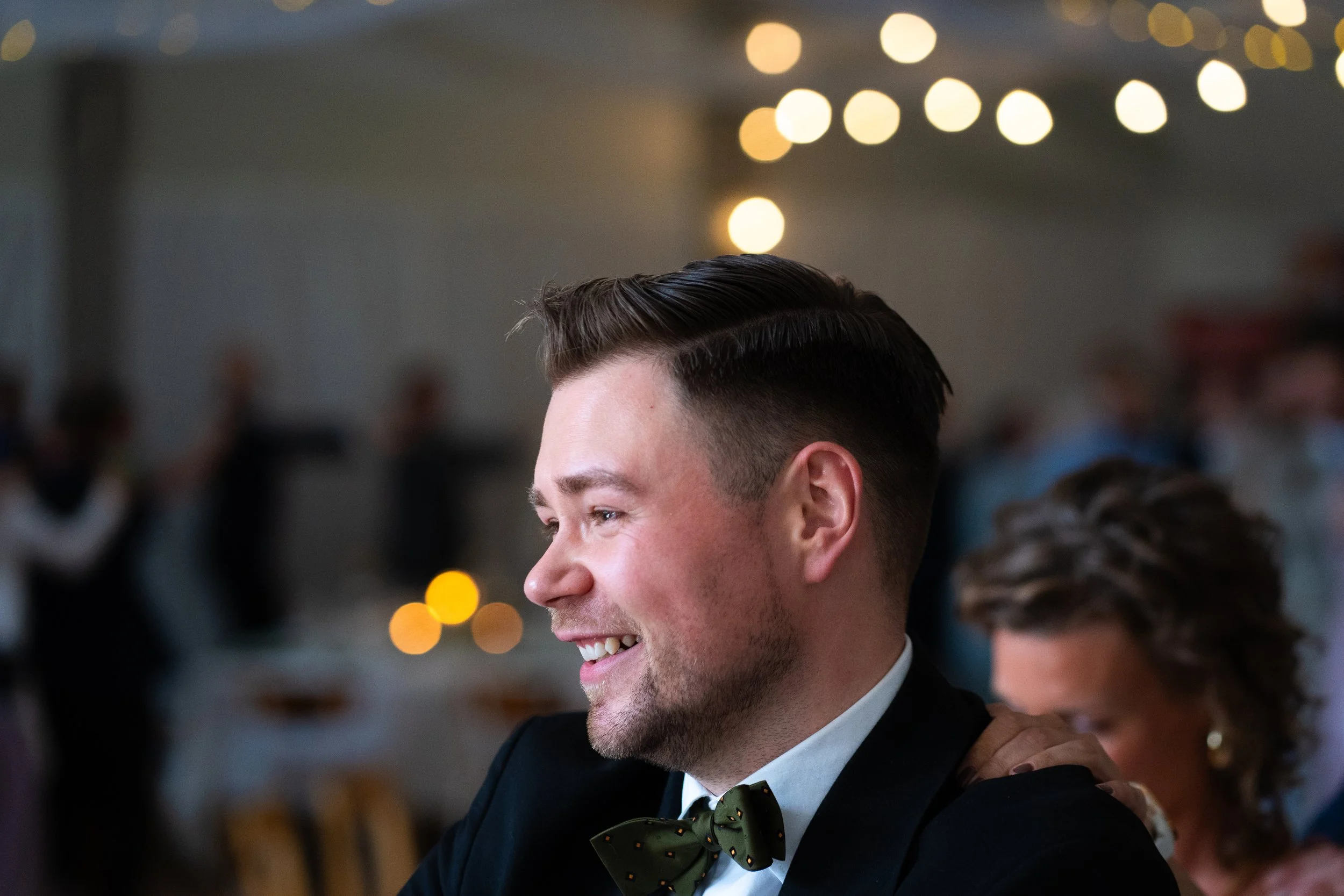 En glad man med mörkt hår i tuxedo och svart fluga vid en fest eller bröllop, med bakgrund av bokeh-ljus och andra gäster.