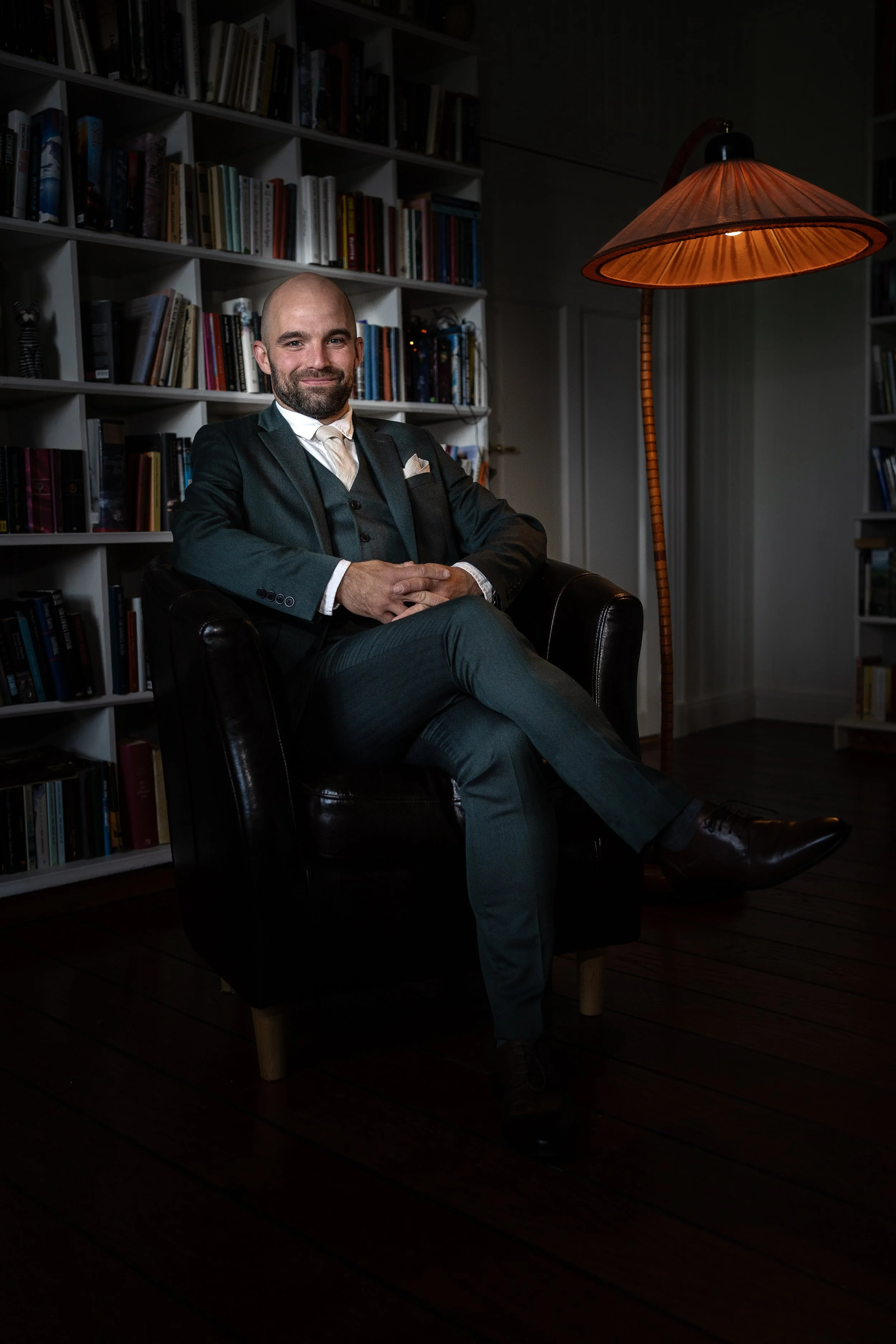 En man i formell kostym som sitter i en fåtölj i ett rum med bokhyllor och en golvlampa.