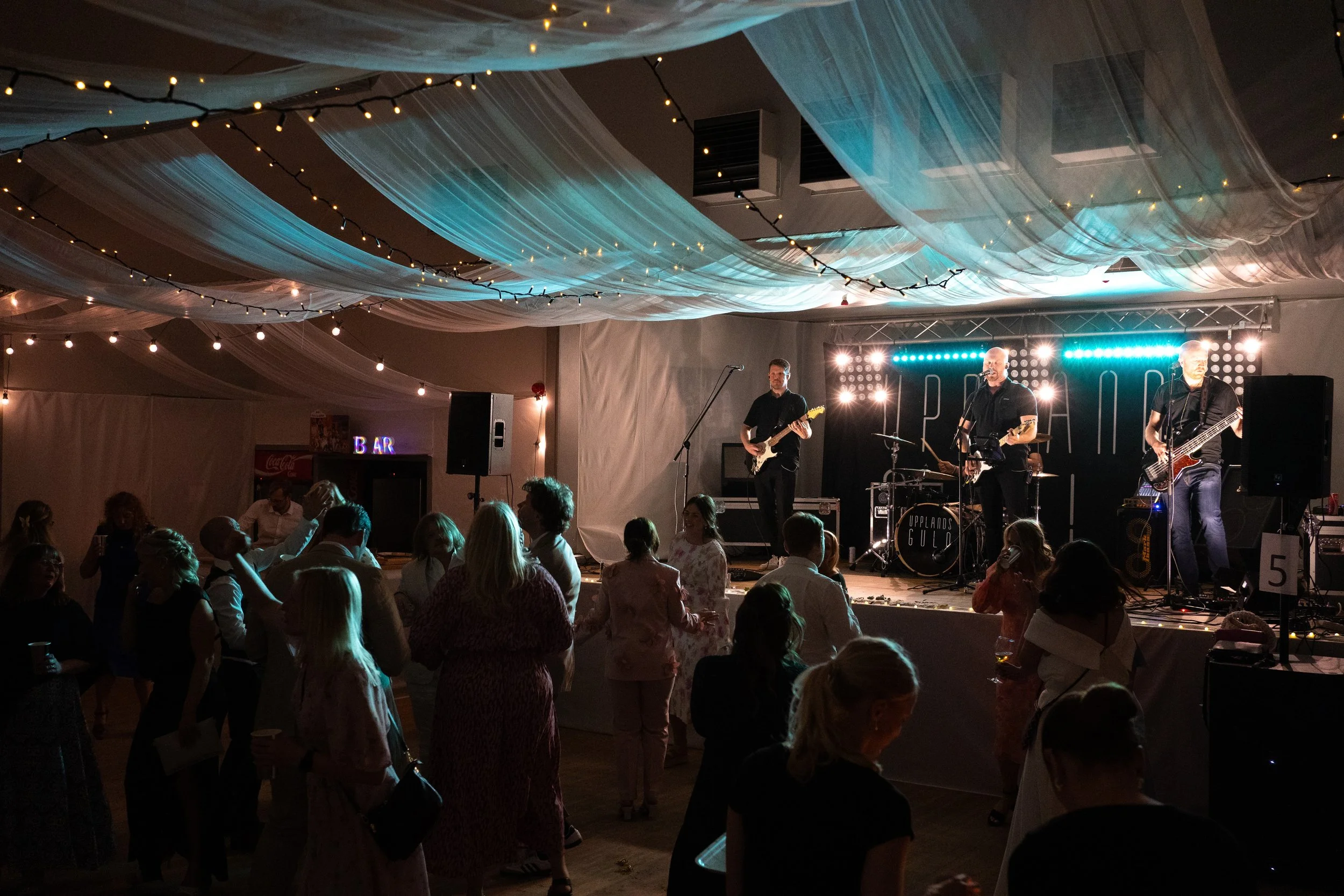 En livespelning med ett band av tre musiker på scenen, publik dansar och njuter, dekoration med ljusslingor och något tyg på taket i en festlokal.