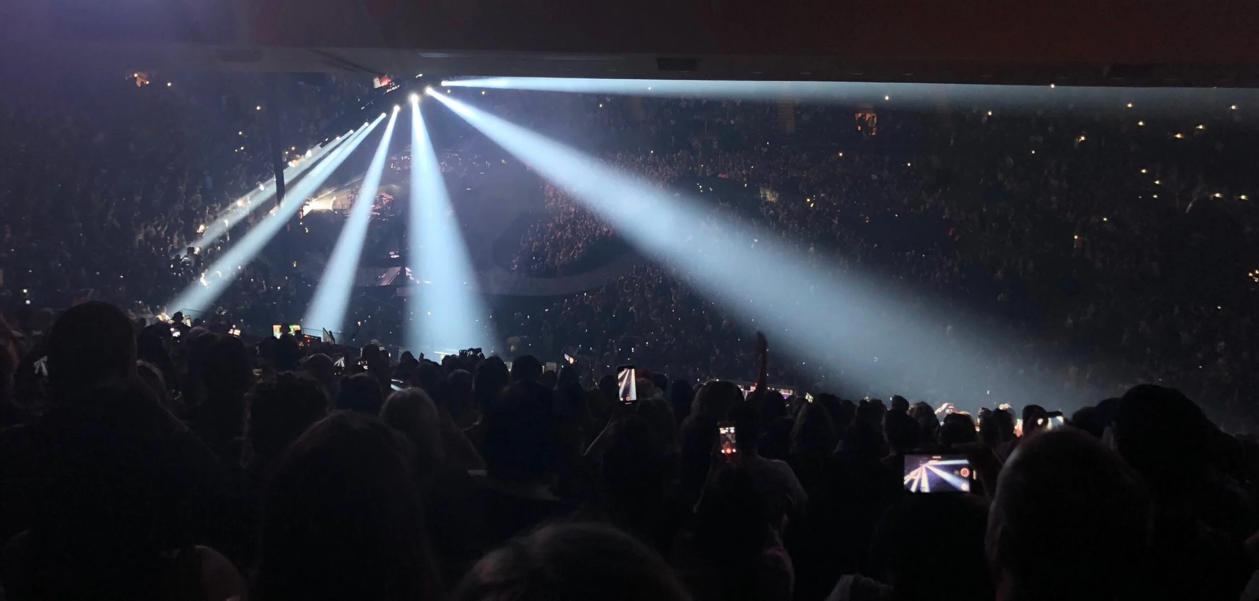 Concert hall filled with audience, bright spotlights shining onto the stage, audience members taking photos.