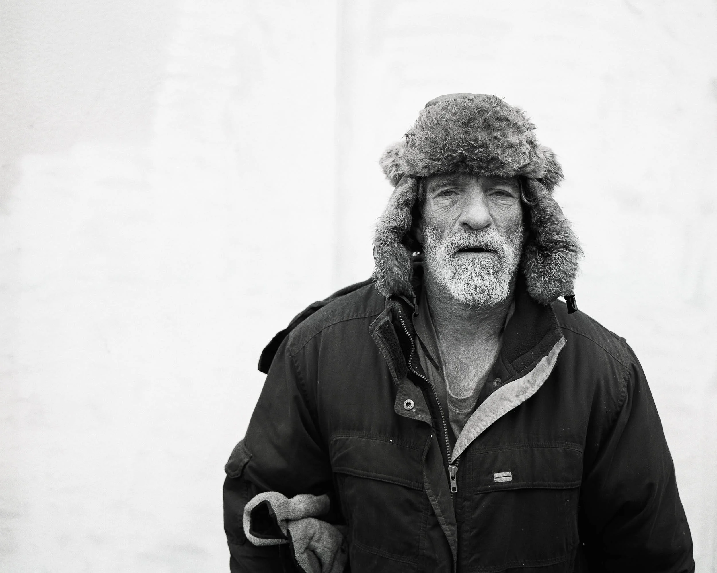 A black and white photo of an elderly man with a beard, wearing a thick furry hat and a dark winter jacket with a collar, standing outdoors against a plain background.