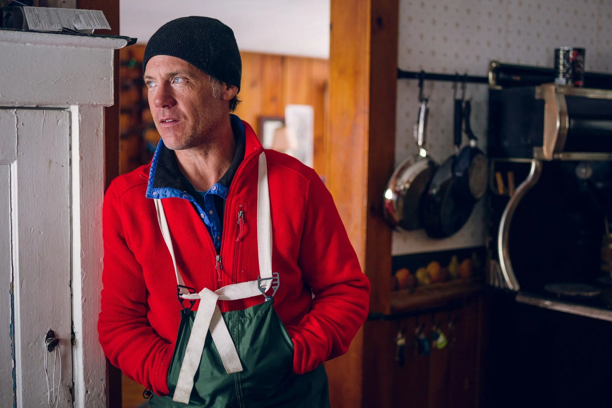 A man wearing a black beanie, red jacket, and apron stands in a cozy room, looking thoughtfully to his left. Behind him are wooden walls, a shelf with a partially visible bicycle wheel, and various household items.