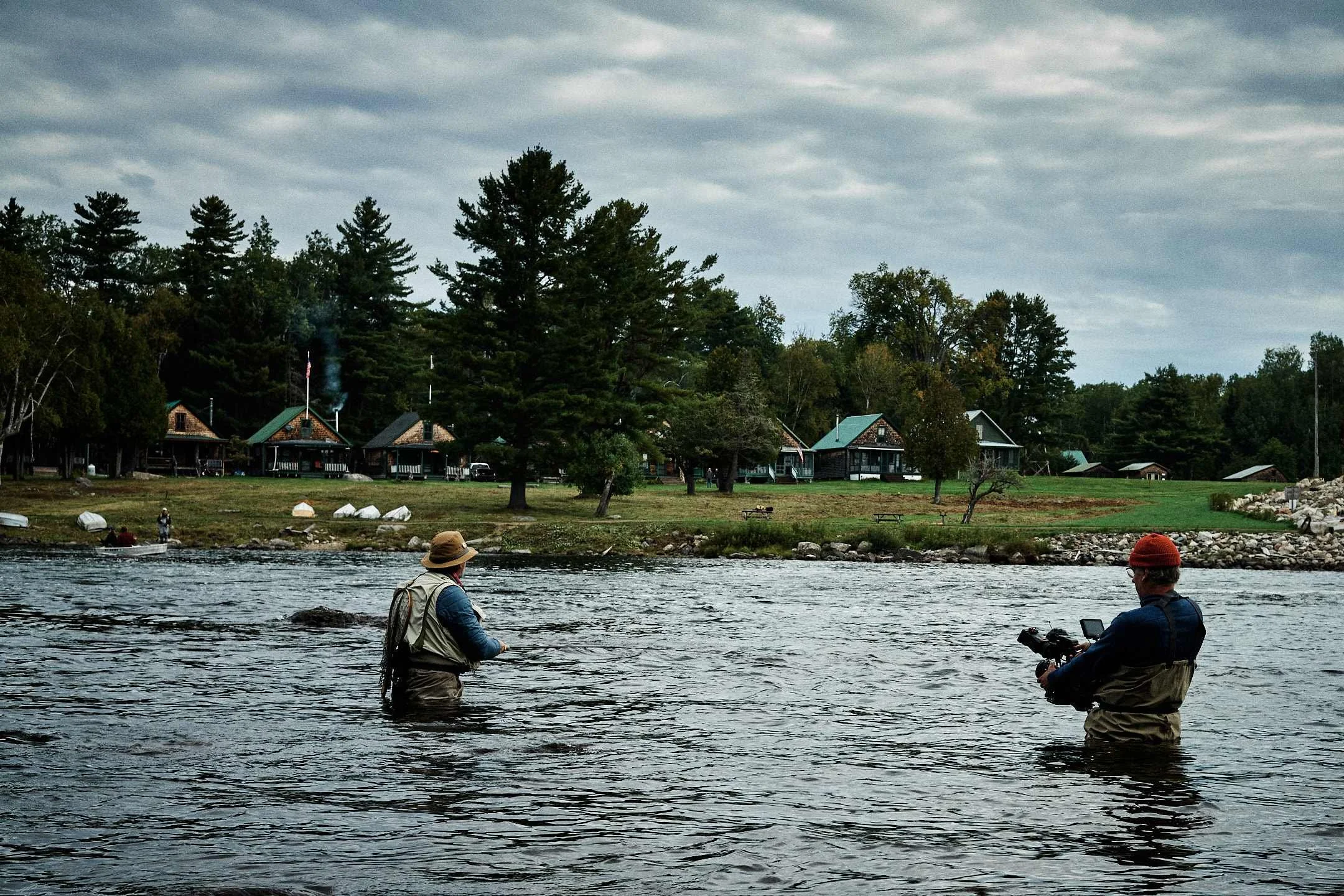 A documentary cameraman in waders filming a fly fisherman in a river with cabins in the background.