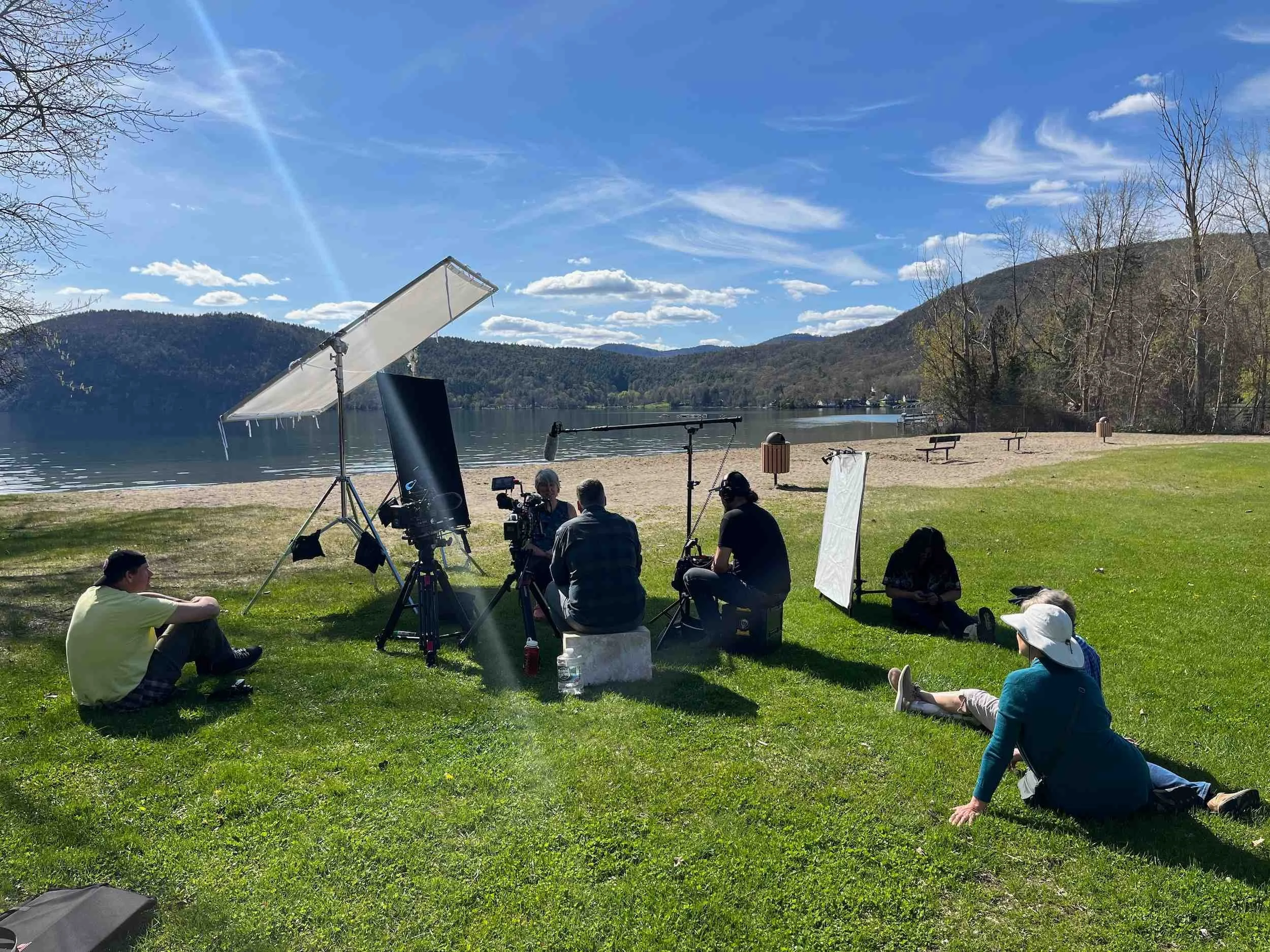 A film crew set up equipment by a lakeside with mountains in the background, filming a scene on a sunny day.
