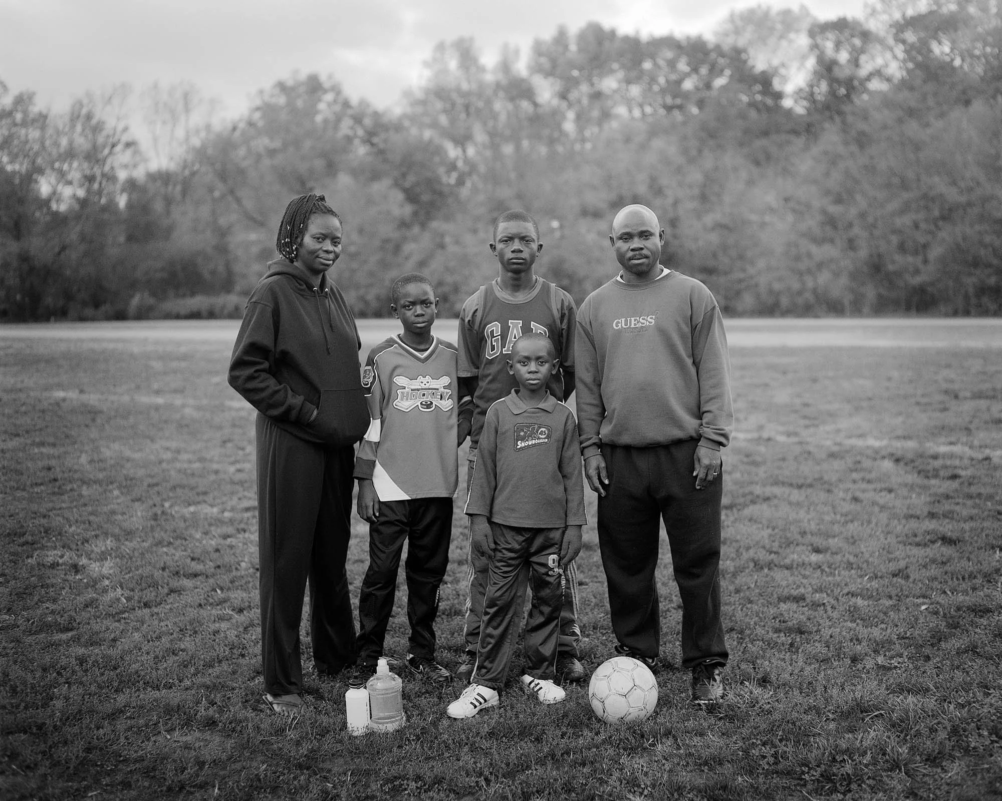 A diverse group of six family members, including two children, standing on a soccer field with a soccer ball and sports bottles in front of them, surrounded by trees in the background, in black and white.