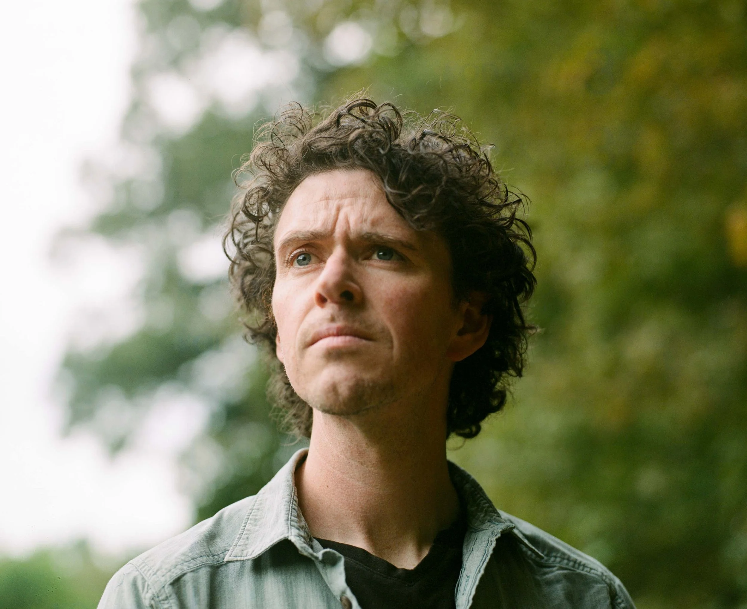 A man with curly dark hair and light skin standing outdoors with a blurry background of trees, wearing a light-colored shirt and black t-shirt, looking thoughtfully into the distance.