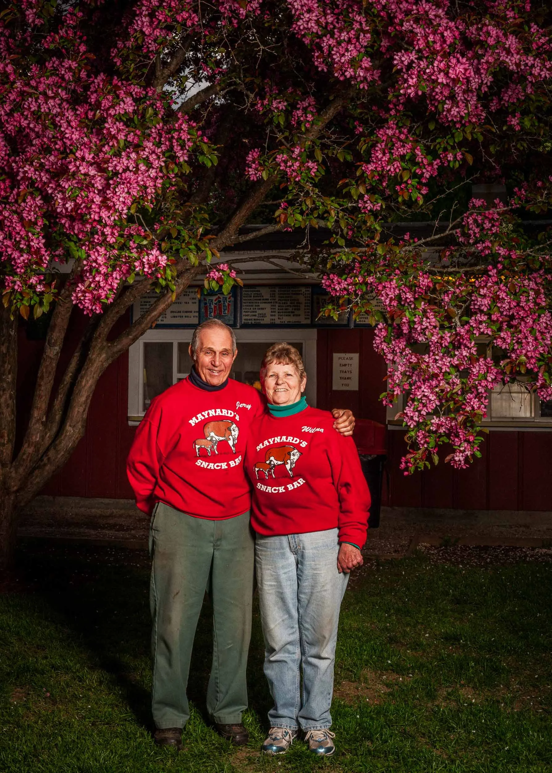 An elderly couple standing outdoors under pink flowering tree, smiling at camera. They are wearing matching red sweatshirts with "Maynard's Snack Bar" logos.