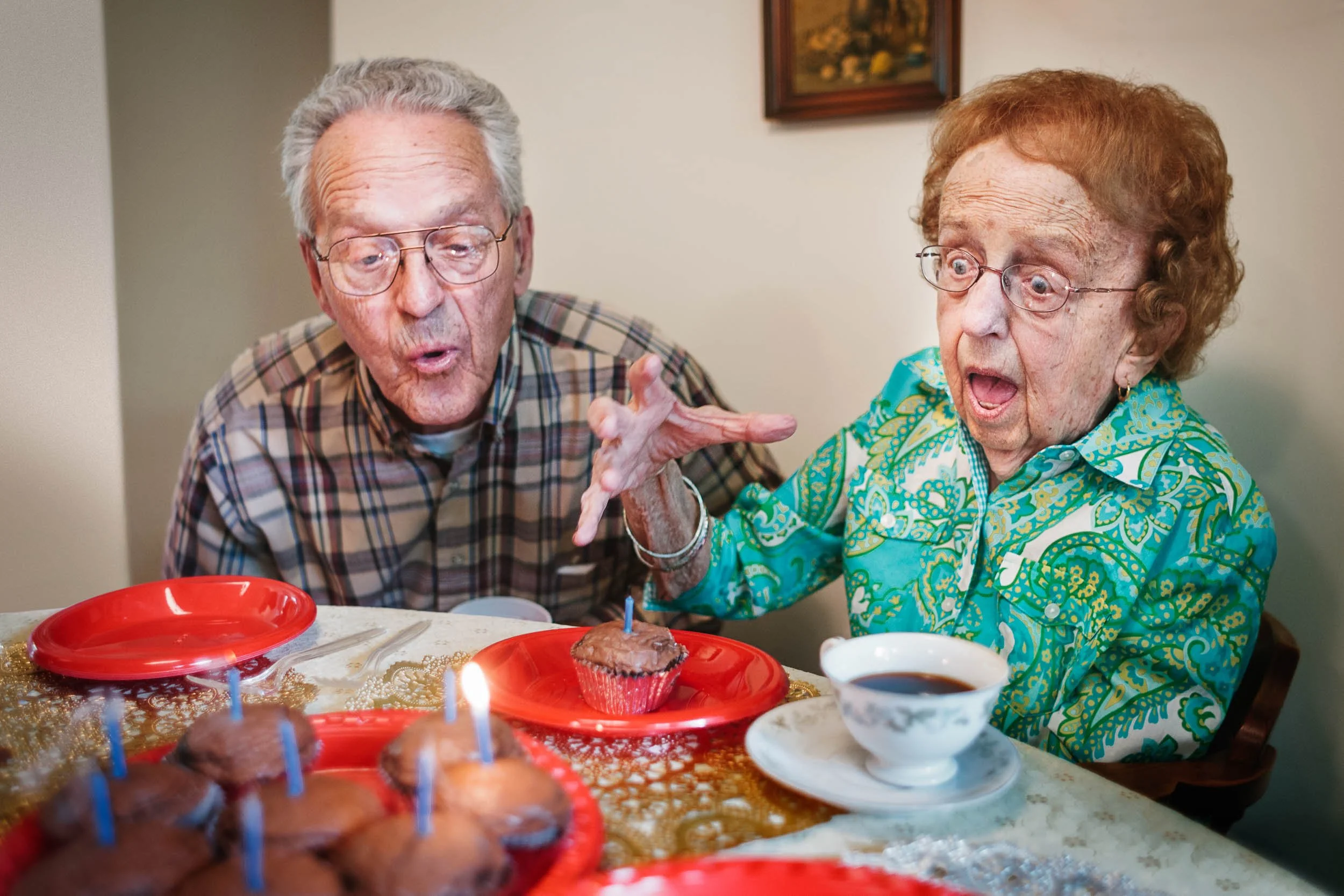 An elderly couple celebrating a birthday at a table with cupcakes, one has a cupcake with a lit candle, both look surprised and happy.