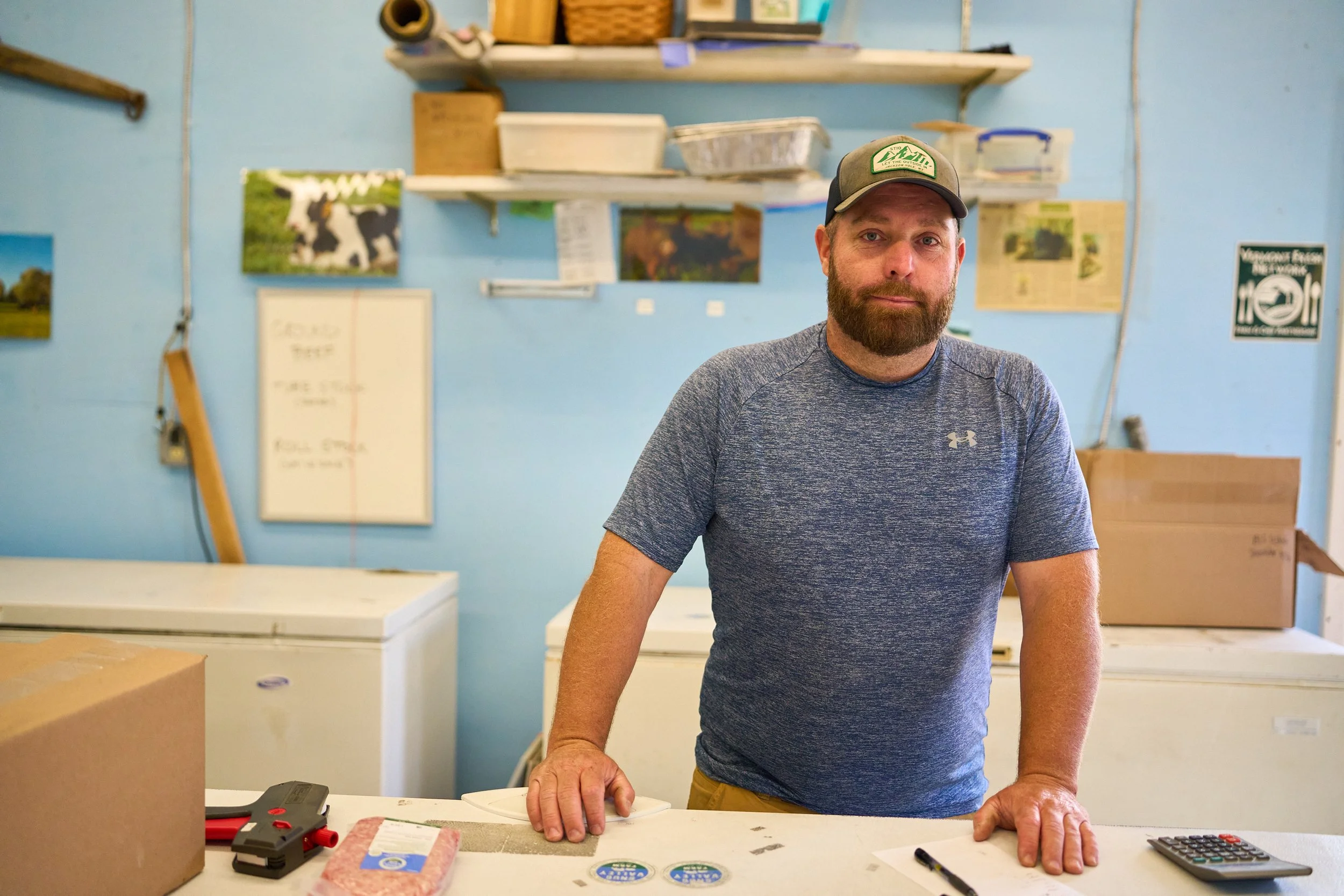 Vermont farmer with a beard wearing a gray Under Armour shirt and a baseball cap standing behind a counter in a store with shelves filled with boxes and pictures of cows on the blue wall behind him.
