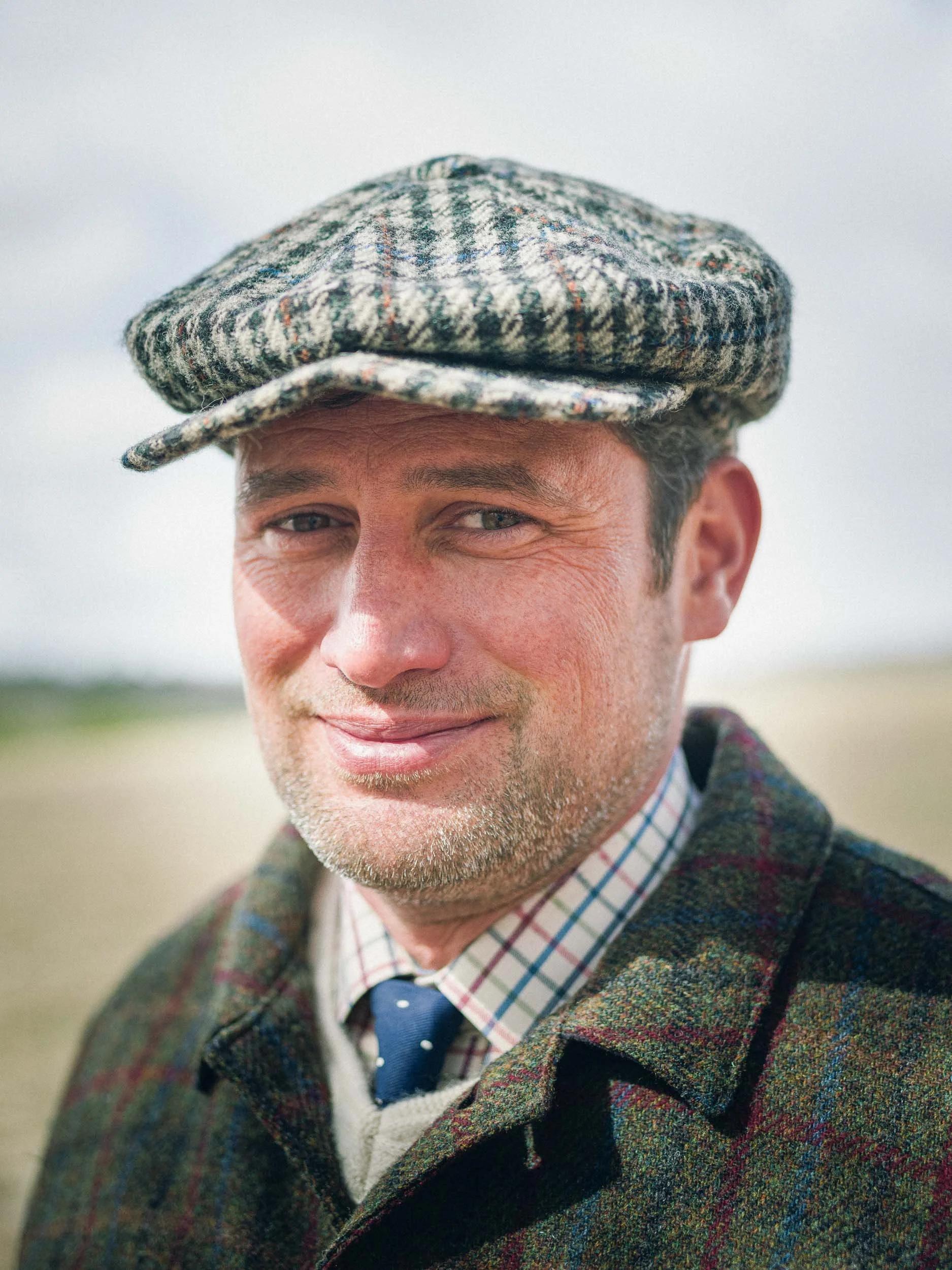 Close-up of a smiling man wearing a gray wool tweed hat, a plaid shirt, a navy tie, and a colorful plaid jacket outdoors on a cloudy day.