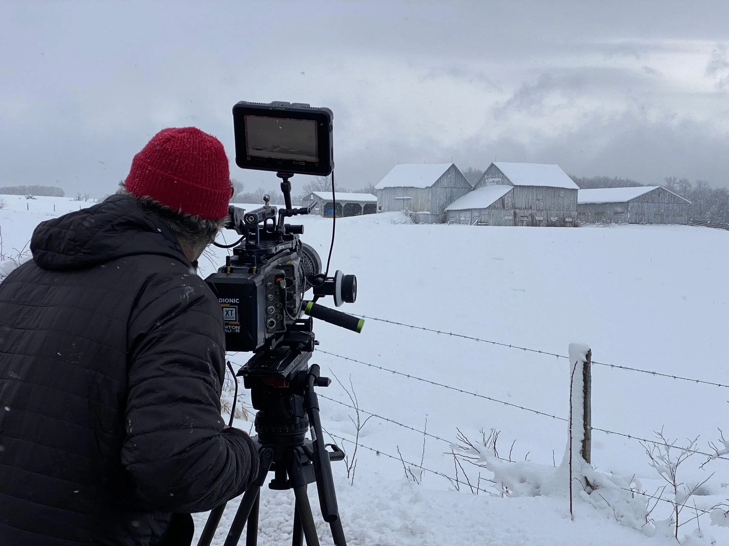 A person in a black jacket and red knit hat operates a professional video camera outdoors in a snowy landscape with a fence and a cluster of barns or farm buildings in the background under a cloudy sky.