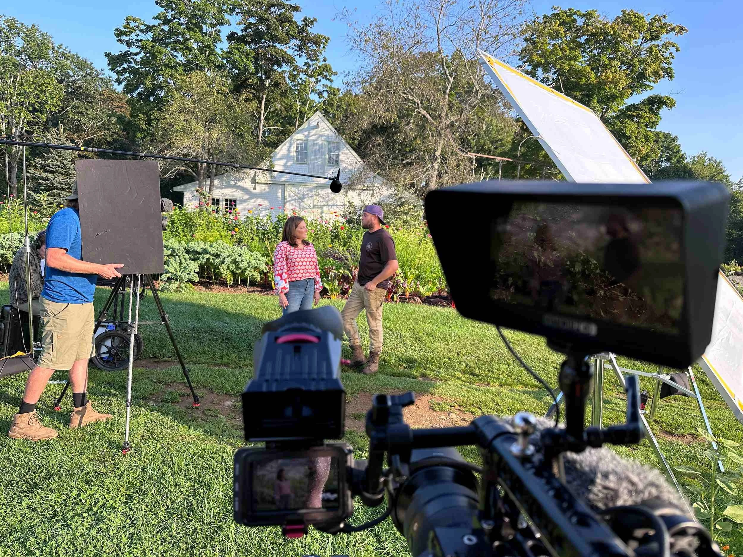 A behind-the-scenes view of a video or photo shoot in a garden, with two people standing and talking, crew and equipment visible including cameras, lights, and reflectors, and a small white house and trees in the background.