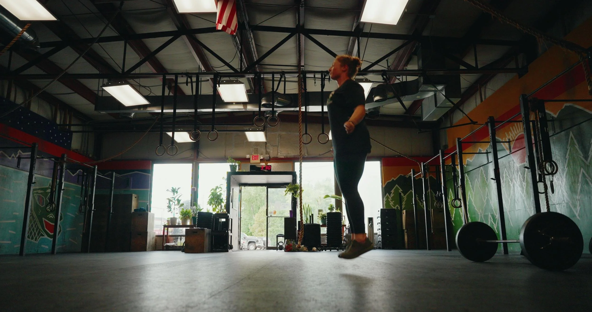 A woman with a bun hairstyle jumping rope inside a gym, with gym equipment and large windows in the background.