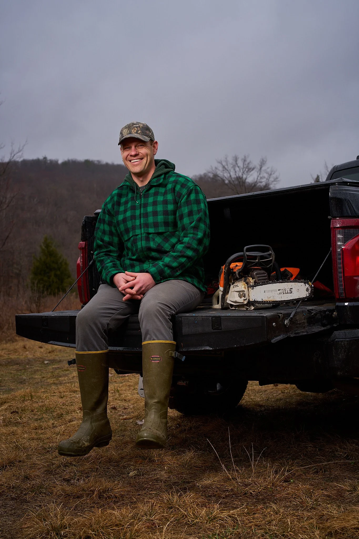 A smiling man sits on the tailgate of a pickup truck, wearing a green plaid jacket, gray pants, camouflage hat, and green rubber boots, outdoors on a cloudy day with bare trees and a hillside in the background.