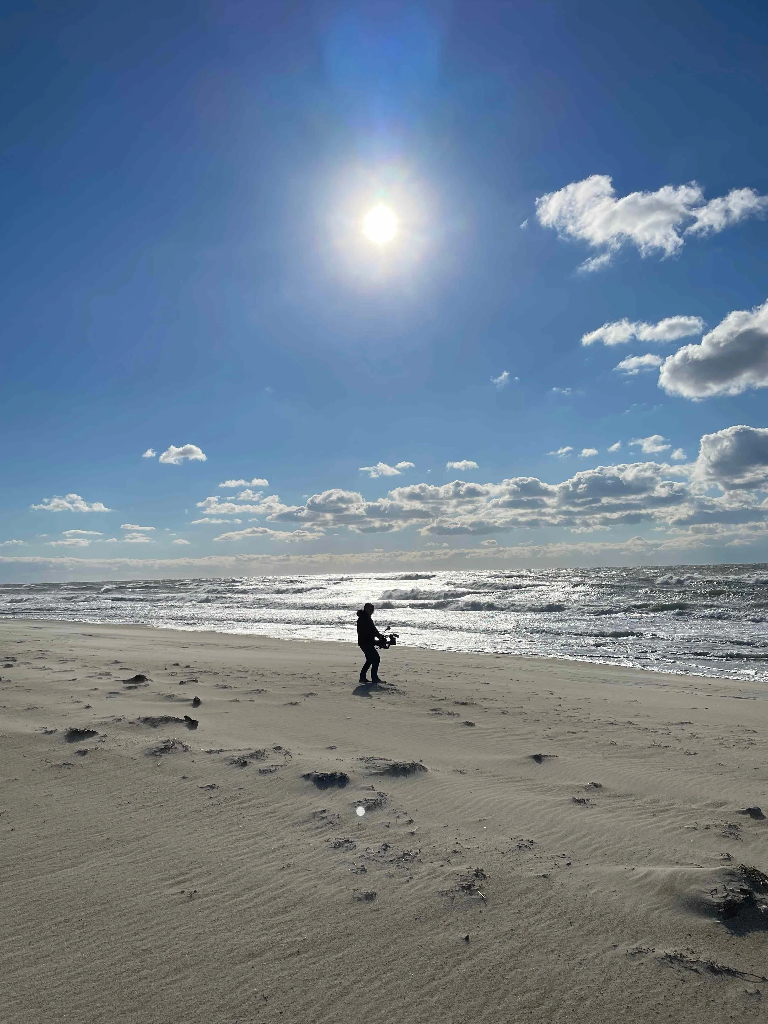A person standing on a sandy beach, possibly with a camera, facing the ocean under a bright sun in a partly cloudy sky.