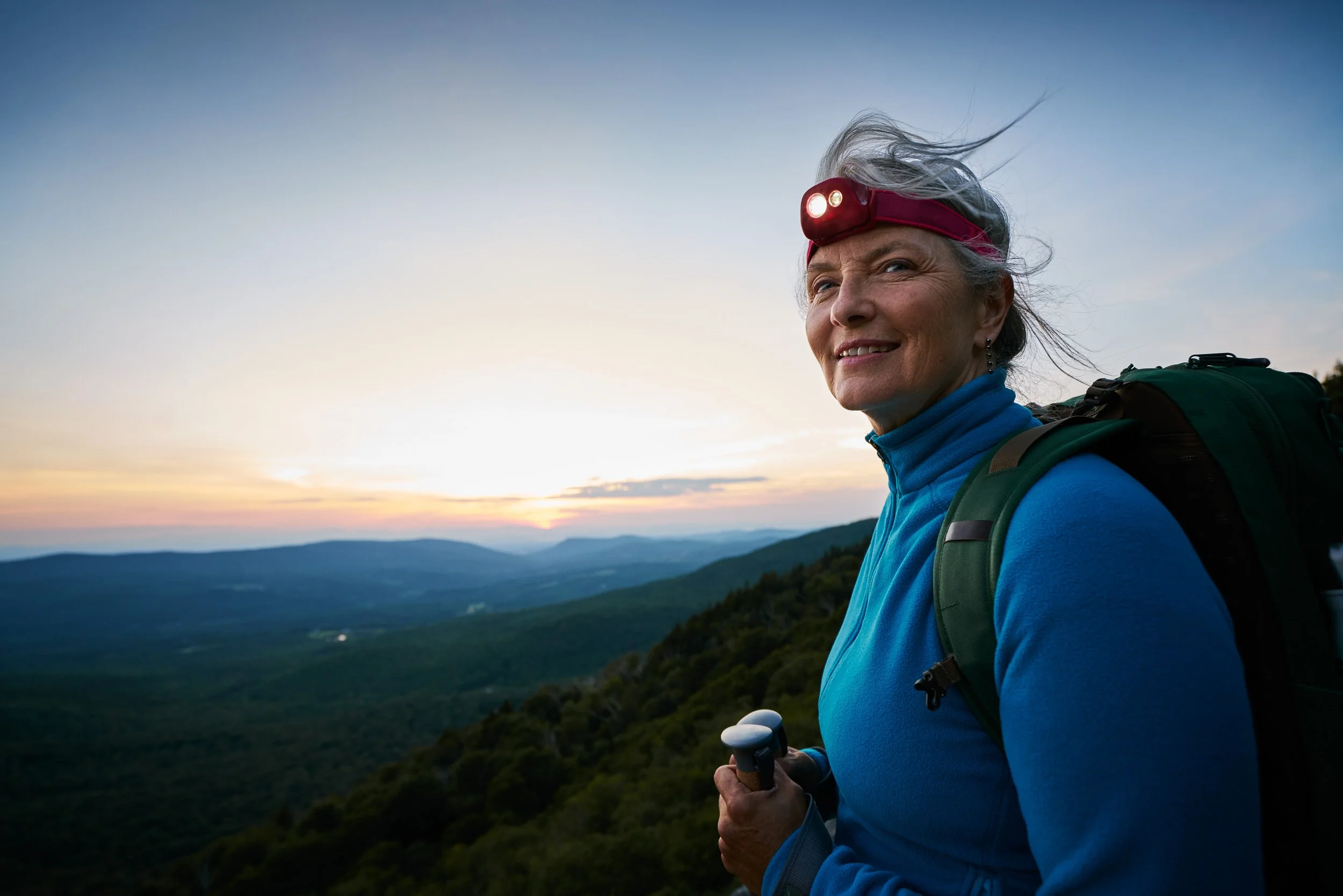 An elderly woman with gray hair, wearing a red headlamp and blue hiking jacket, carrying a backpack and trekking poles, enjoying a scenic mountain view at sunset.