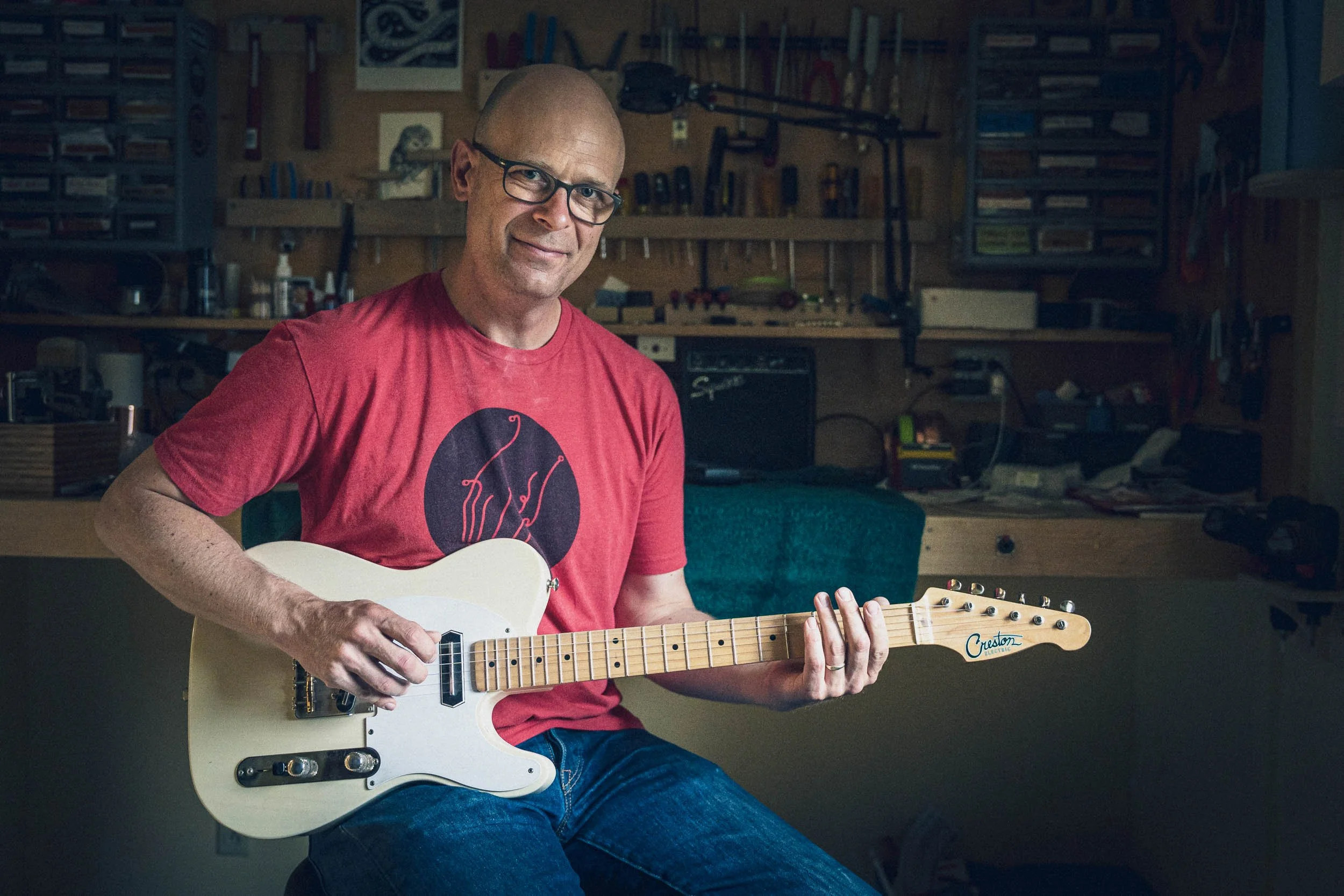 A middle-aged man with glasses, wearing a red t-shirt with a tent graphic, playing an electric guitar in a woodworking or crafting workshop with shelves, tools, and artwork in the background.