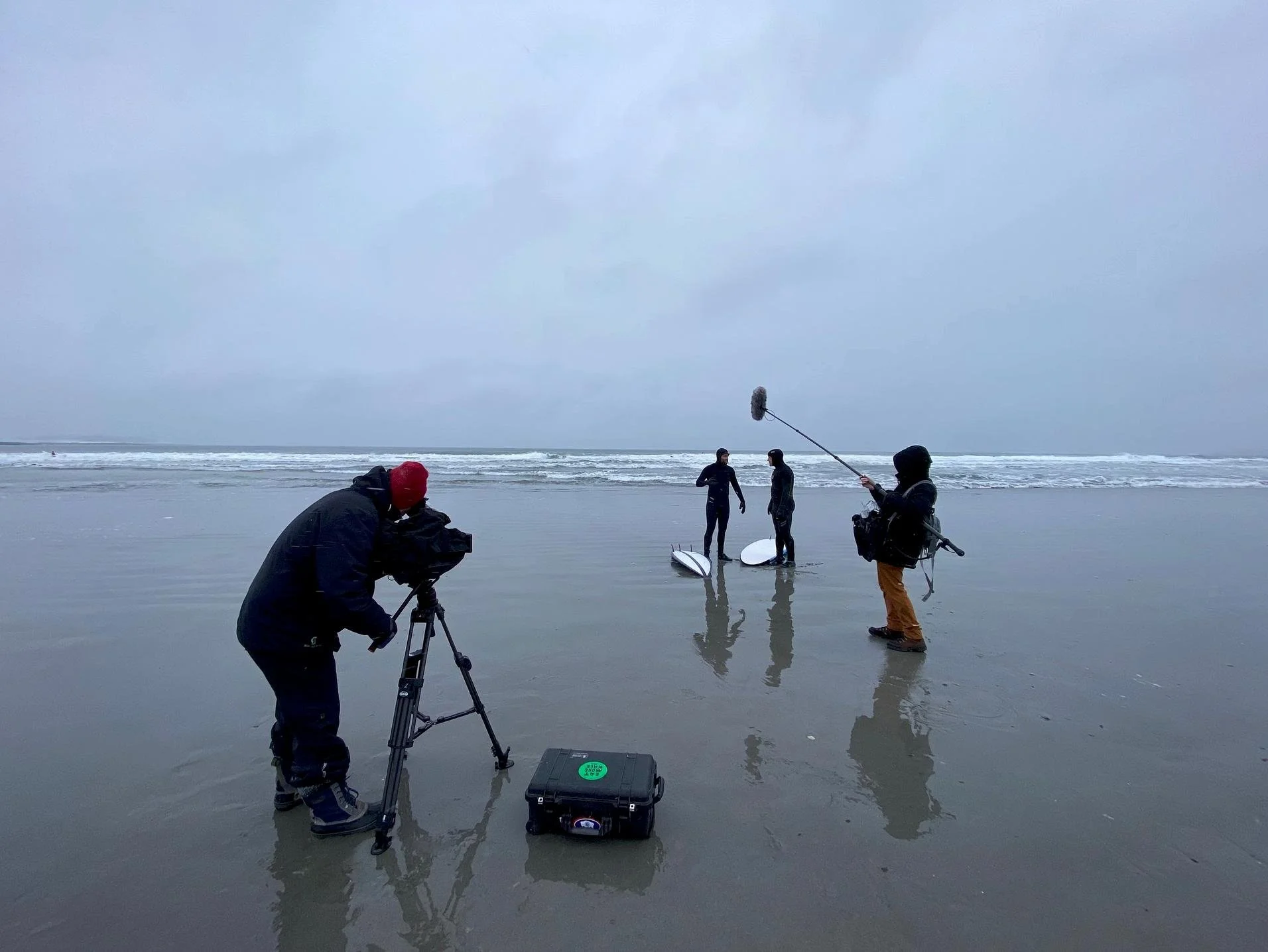 Film crew filming on a cloudy beach with three people standing near surfboards and a photographer capturing the scene.