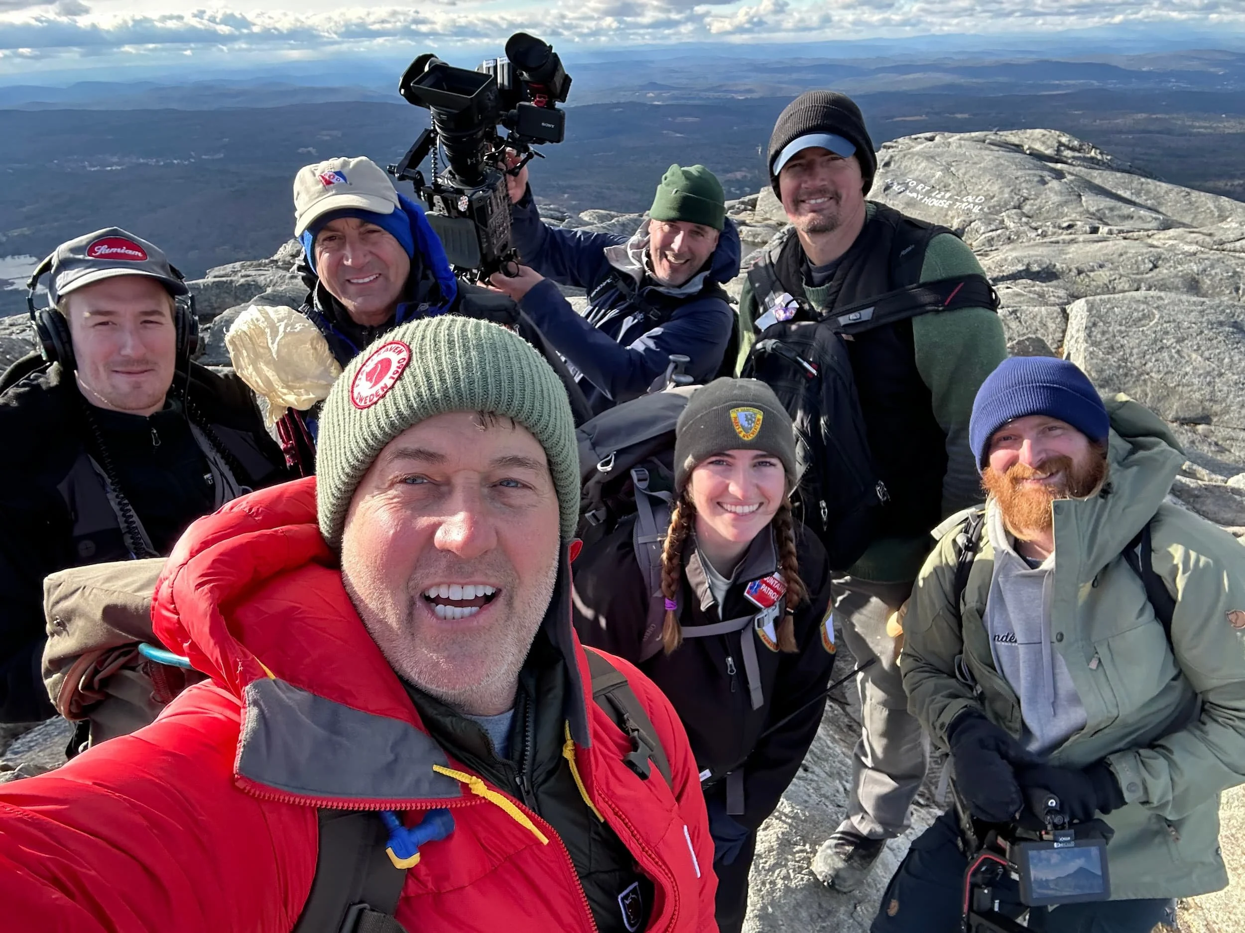 A group of people on a mountain summit taking a selfie with a scenic view of mountains and sky in the background.