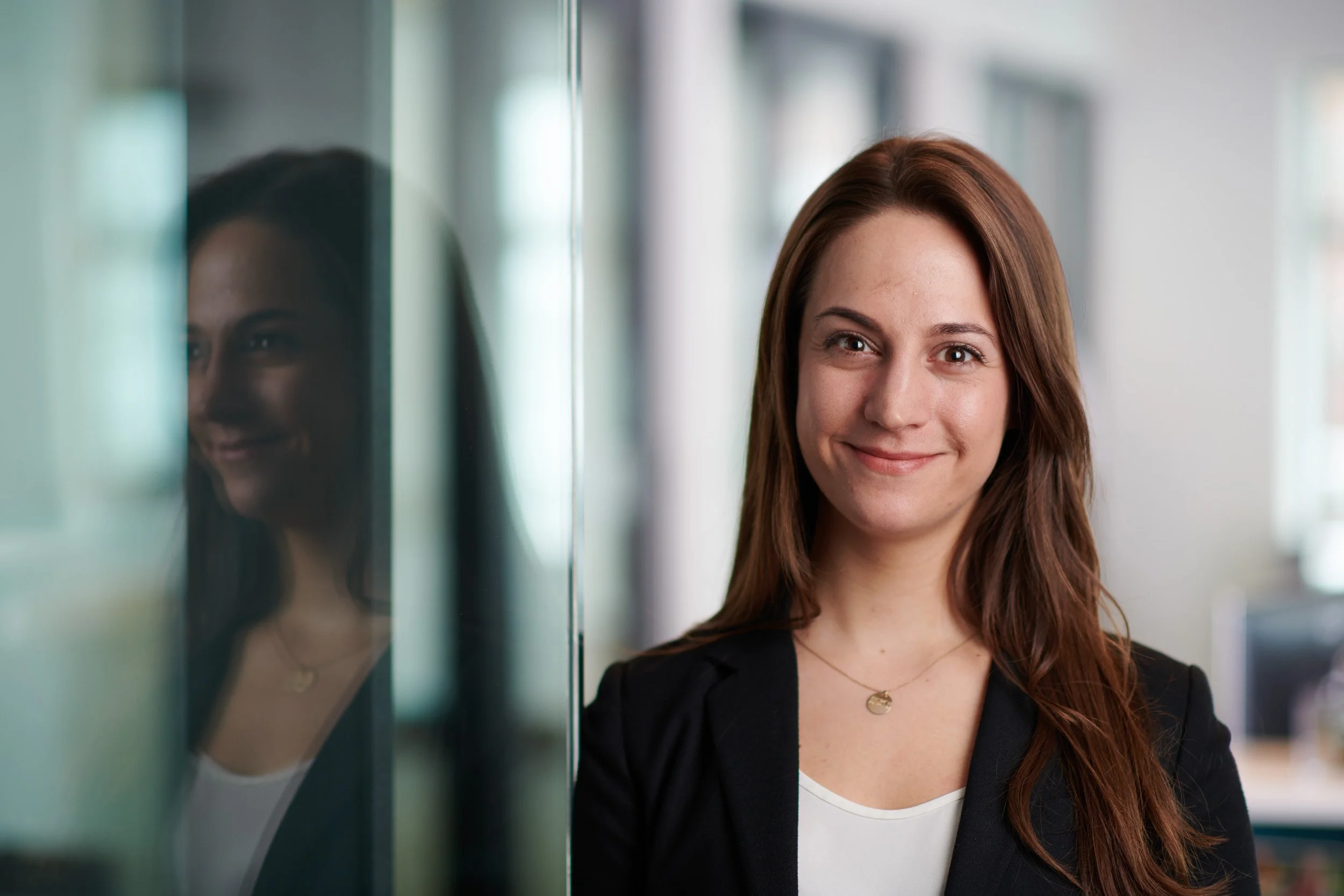 A woman with long brown hair smiling, standing next to a reflective glass surface in a modern indoor setting.