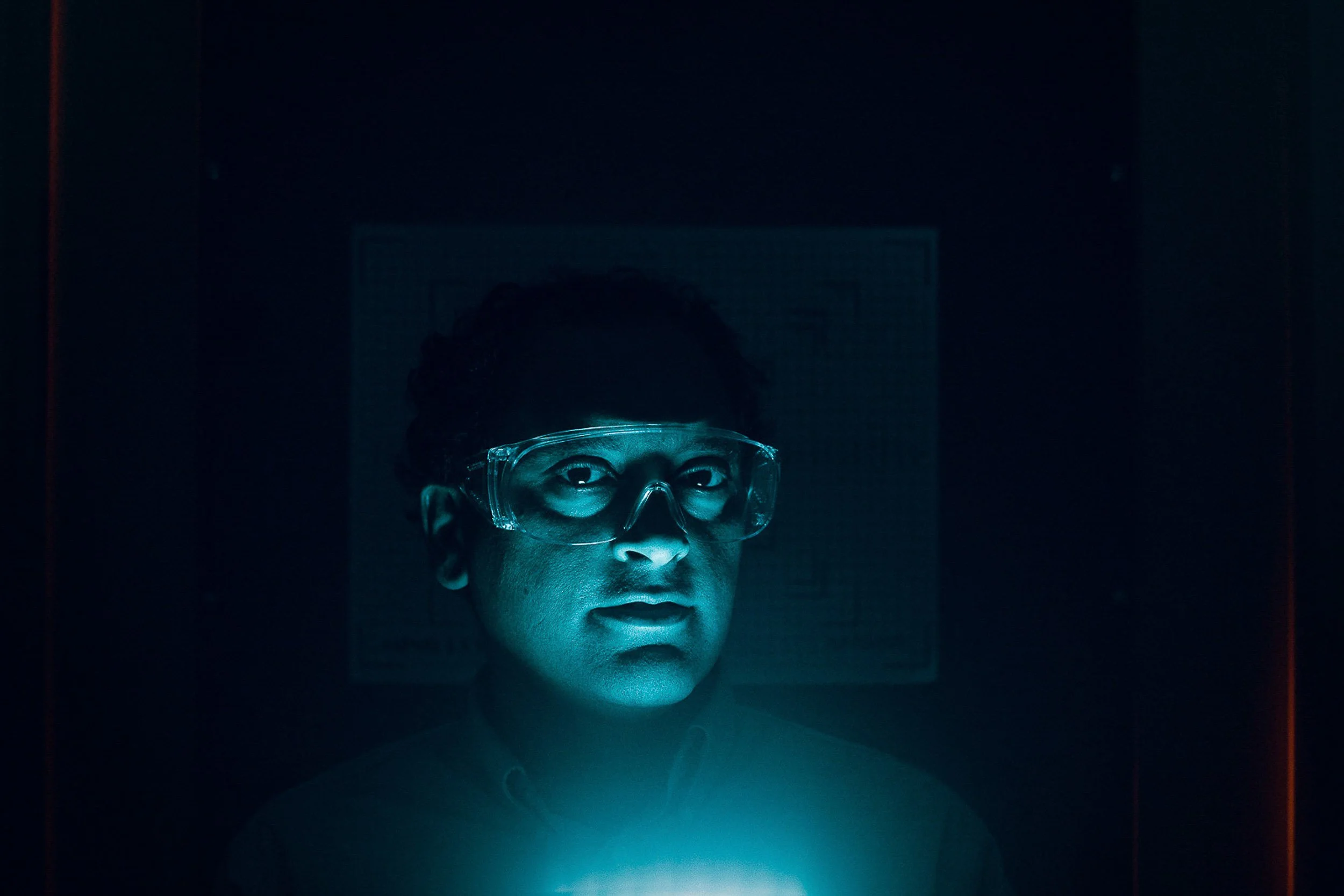 A person wearing safety goggles looking at a screen with a serious expression, illuminated by a blue light in a dark environment.