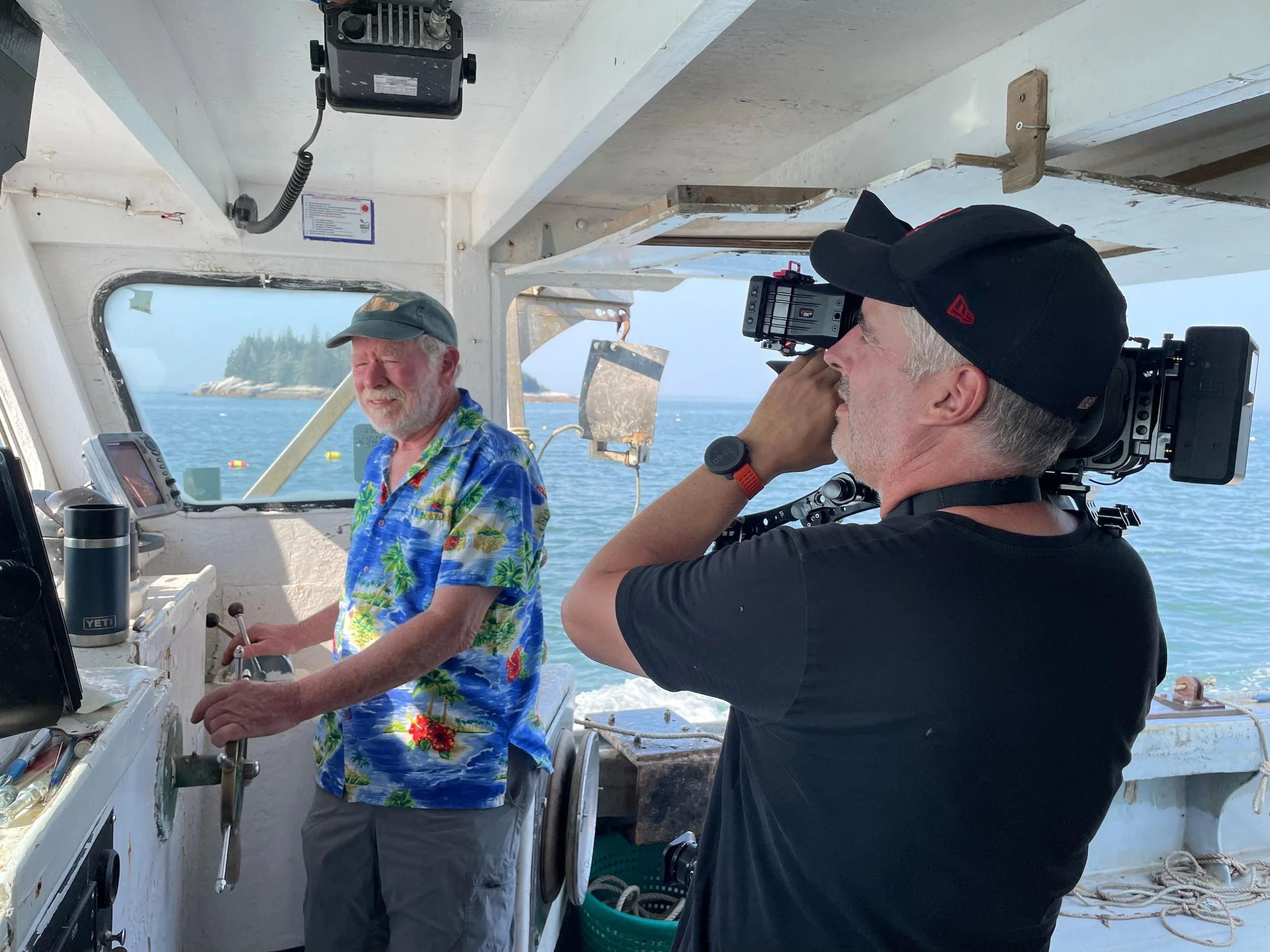Two men on a boat, one with white hair and beard wearing a Hawaiian shirt and cap, the other with gray hair in a black shirt and cap, operating a camera or filming equipment near a control panel with a window showing the ocean and distant shoreline.