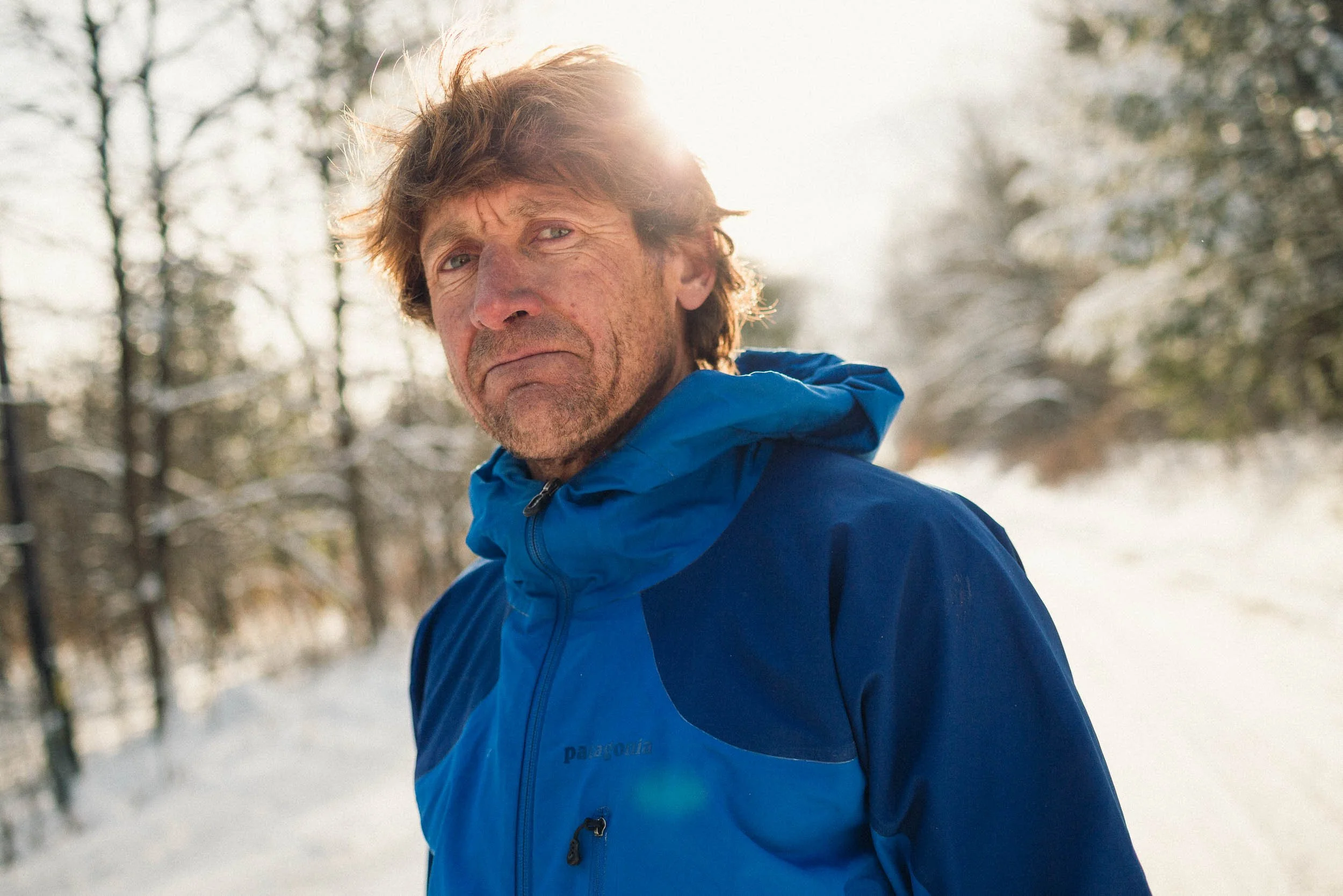 A middle-aged man with tousled brown hair and blue eyes, wearing a bright blue Patagonia jacket, standing outdoors in a snowy forested area with leafless trees, sunlight filtering through the branches.