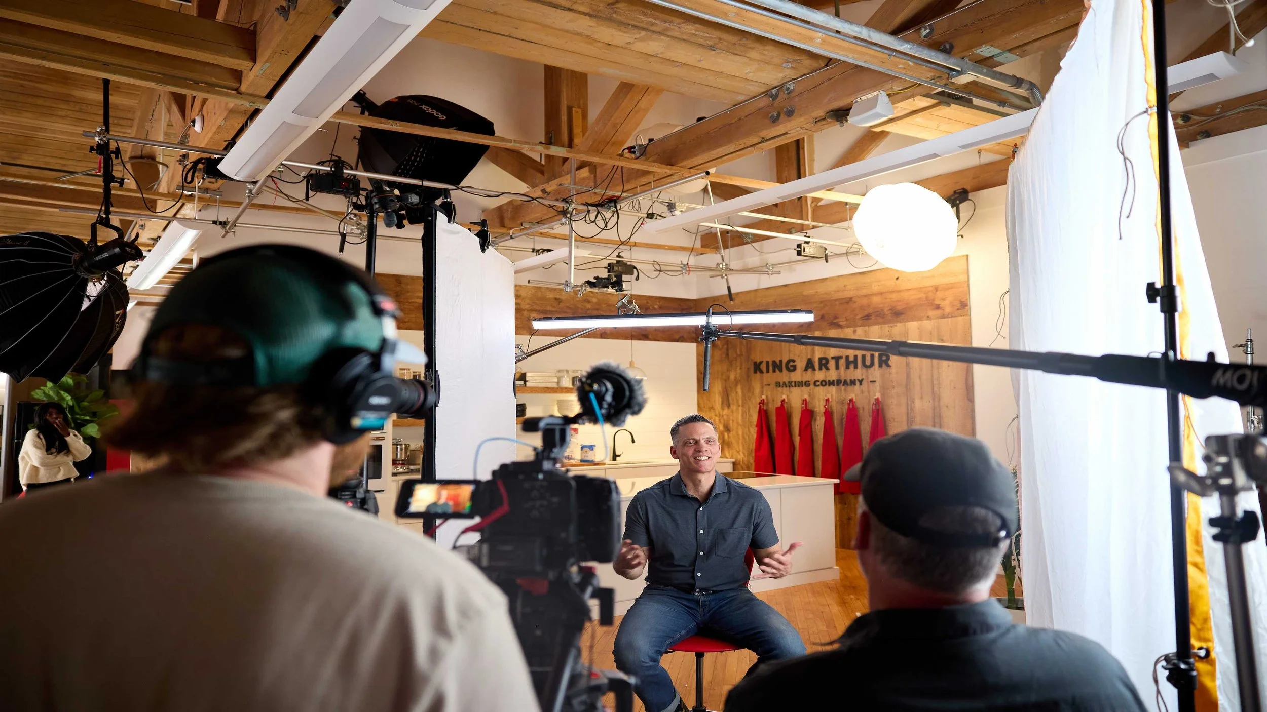 A man seated in front of a camera and crew, speaking during a video shoot or interview in a studio with wooden beams and a sign that says 'King Arthur Baking Company' in the background.