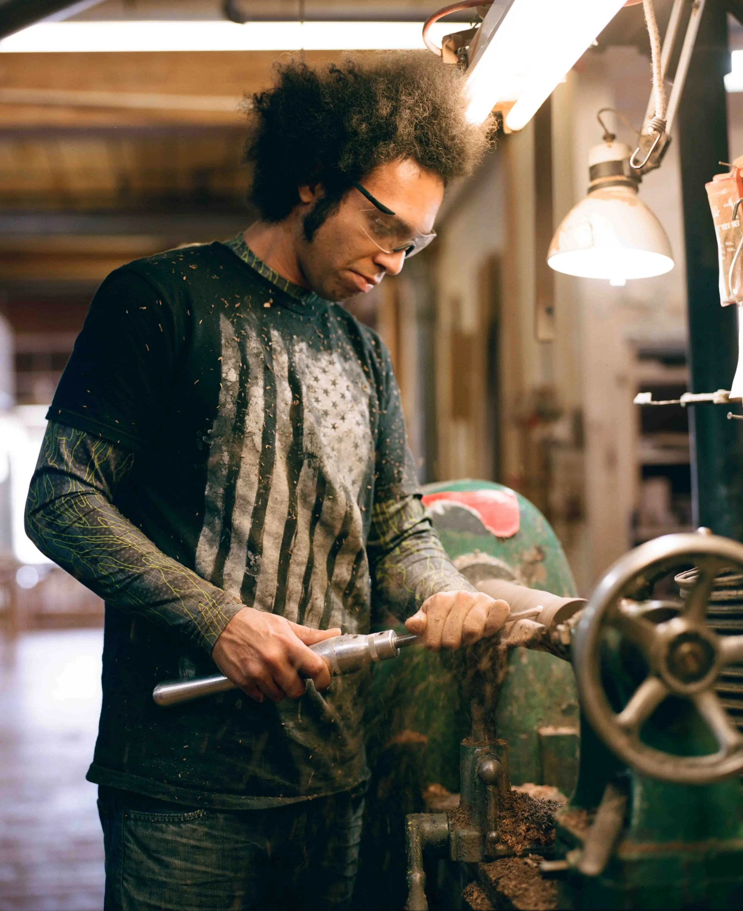 A person with curly hair and safety glasses working with machinery in a workshop environment.